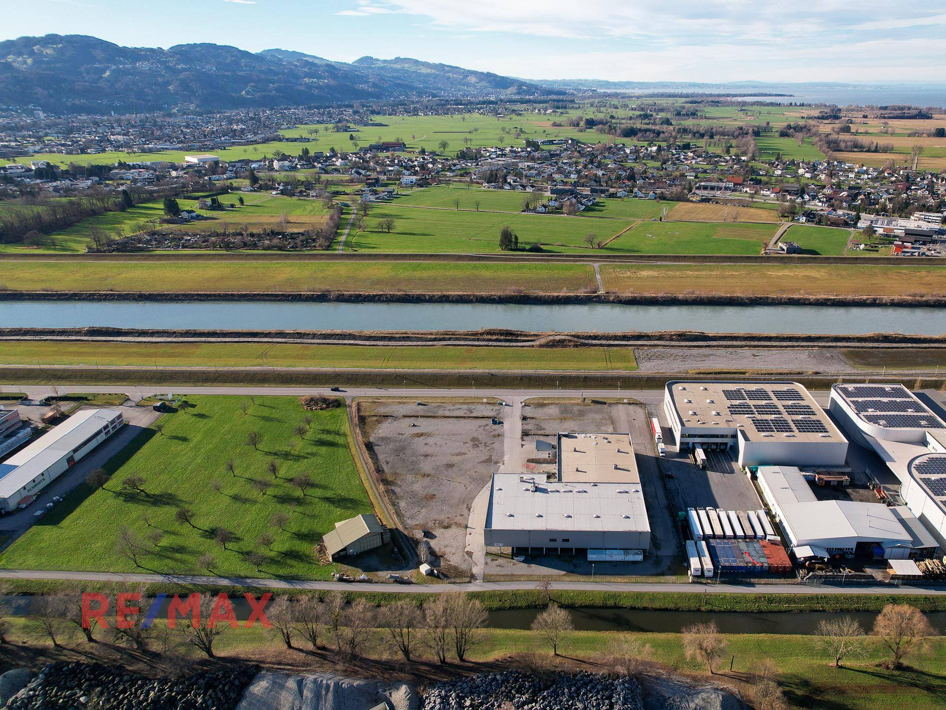 Luftaufnahme des Gewerbegebiets mit grünen Wiesen, einem Kanal und einer beeindruckenden Bergkulisse.