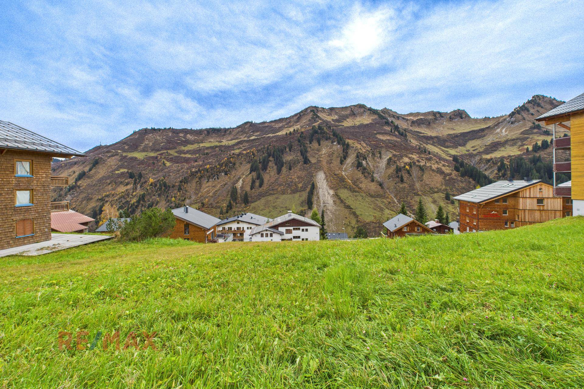 Weite grüne Wiese mit Gebäuden und einer beeindruckenden Bergkette am Horizont.