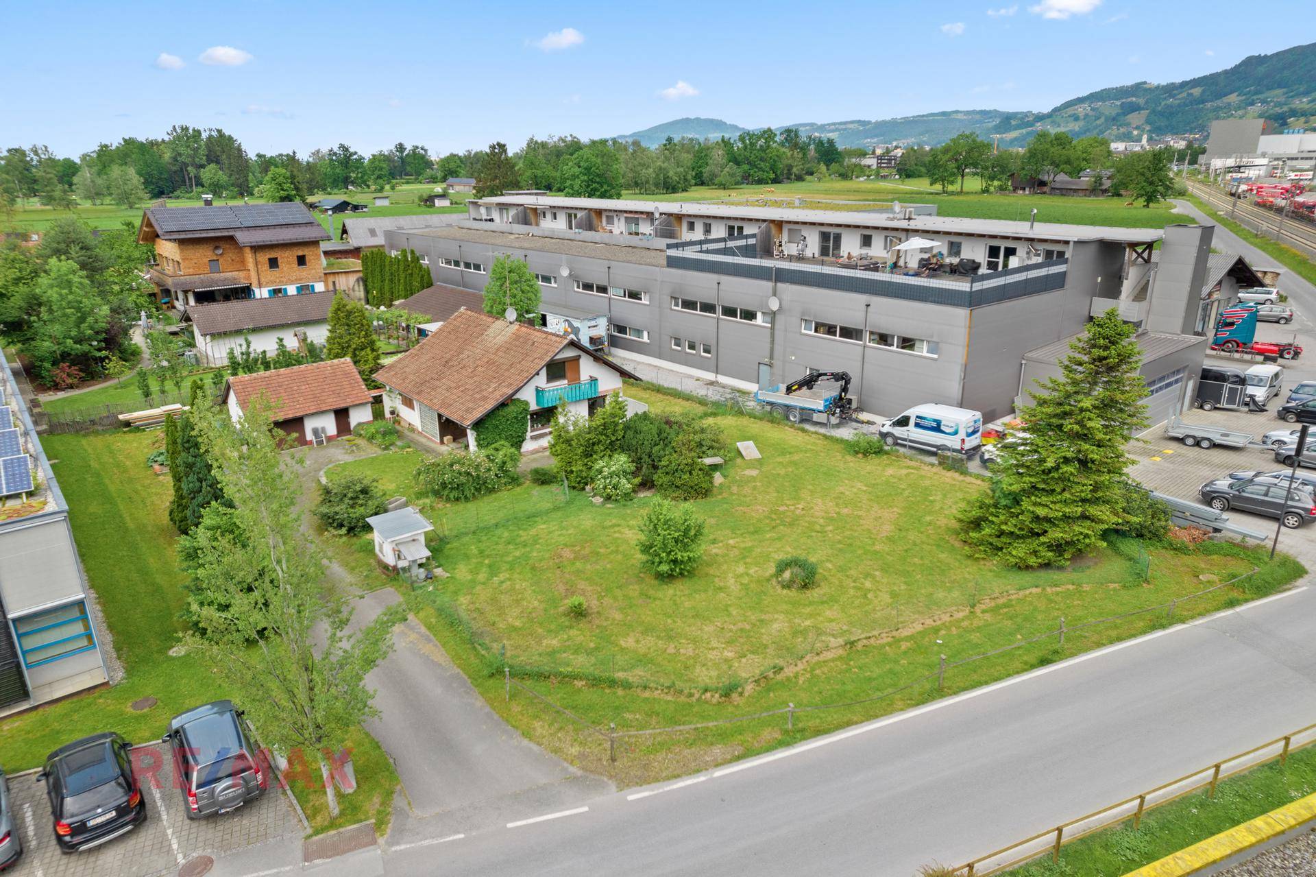 Übersichtsaufnahme des Hauses mit Garten, umgeben von Gewerbebauten, Feldern und Bergen im Hintergrund.