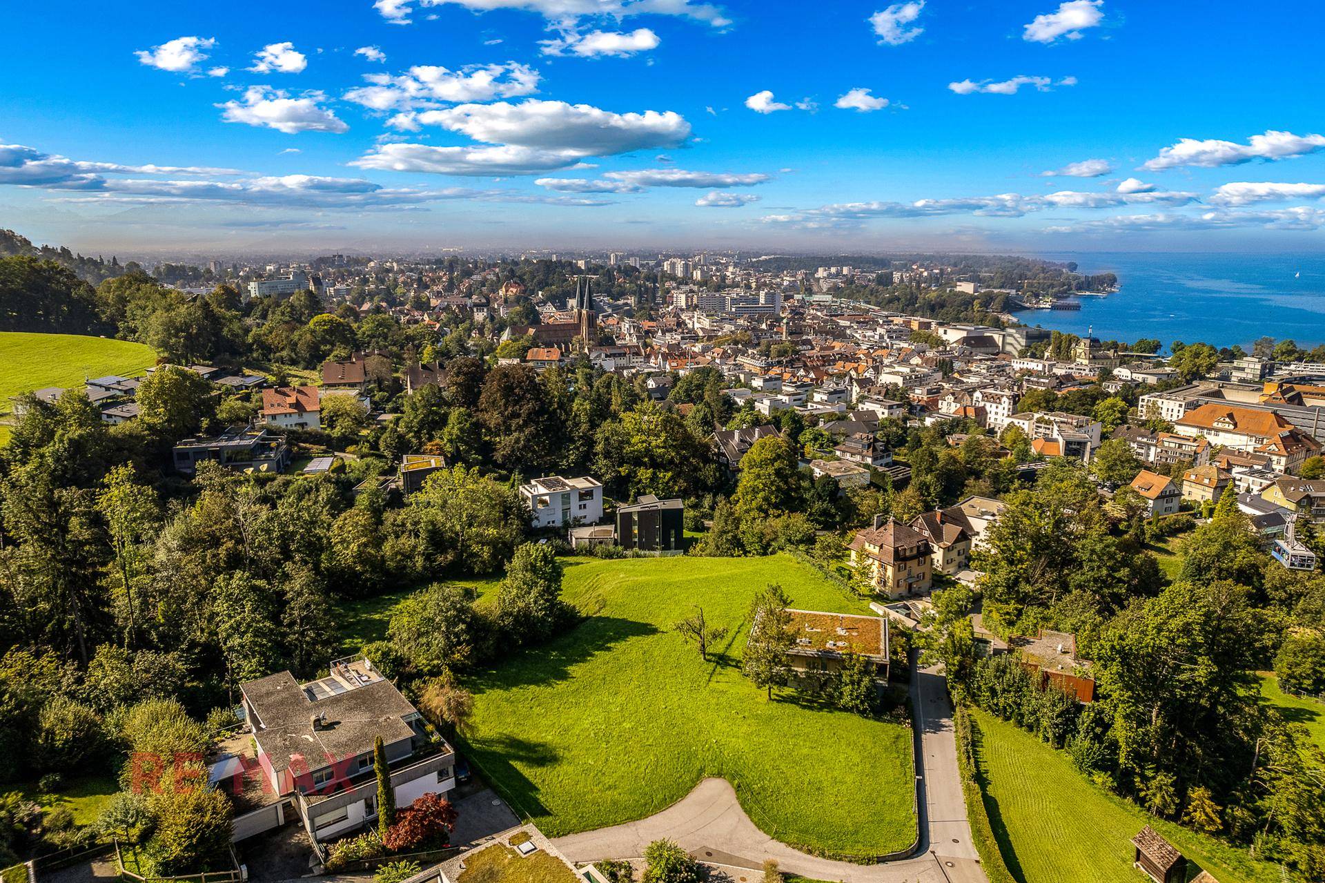 Panoramablick auf eine Stadt am See, umgeben von grünen Hügeln und Wohngebieten.