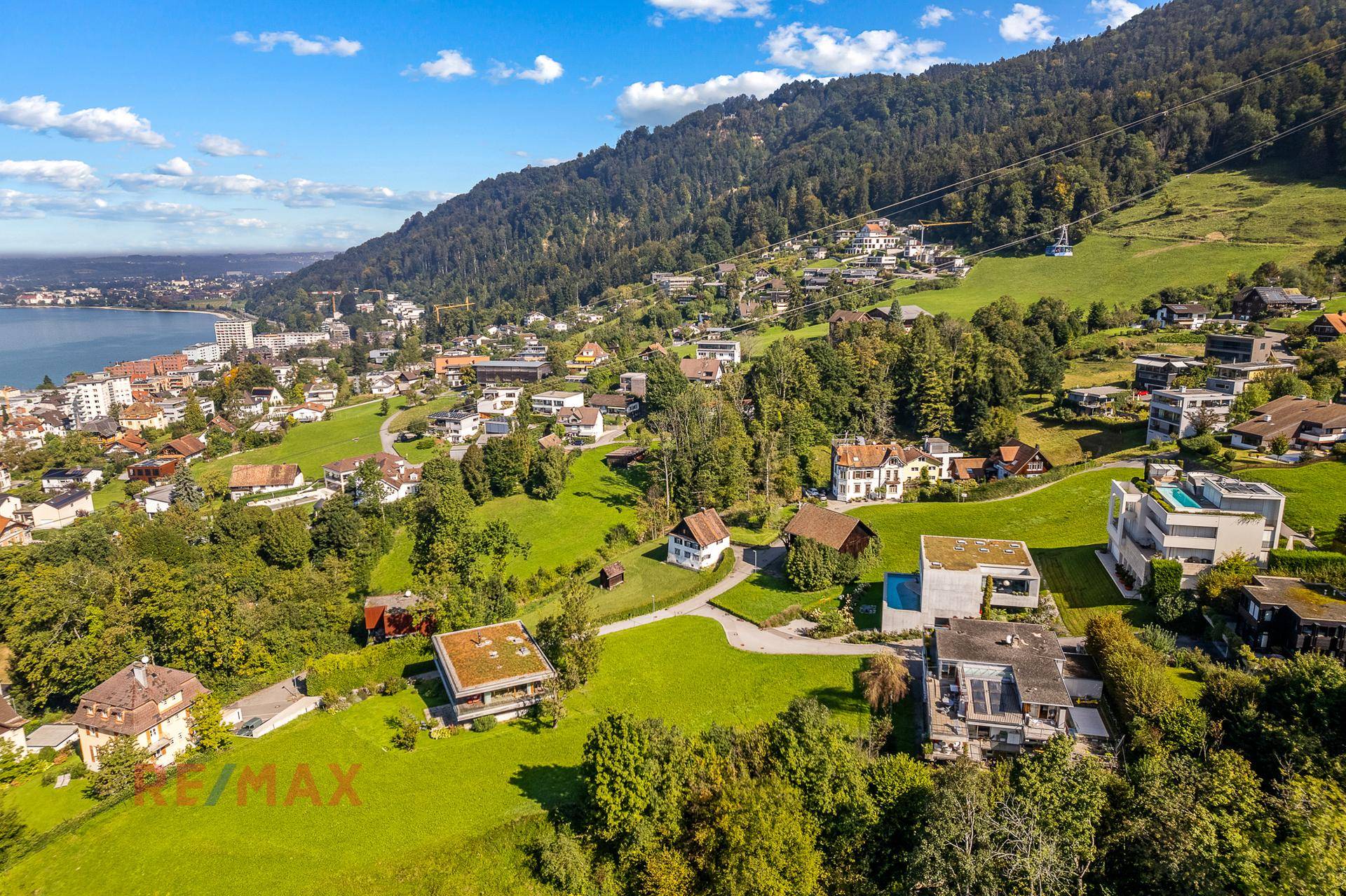 Luftaufnahme einer Siedlung am Hang mit Blick auf den See und die umliegenden Berge.