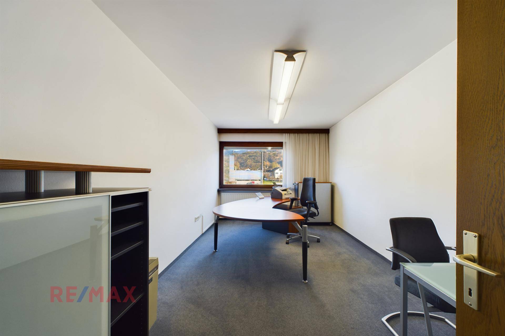 Helles Büro mit Teppichboden, Schreibtisch und Fenster mit Blick ins Grüne.