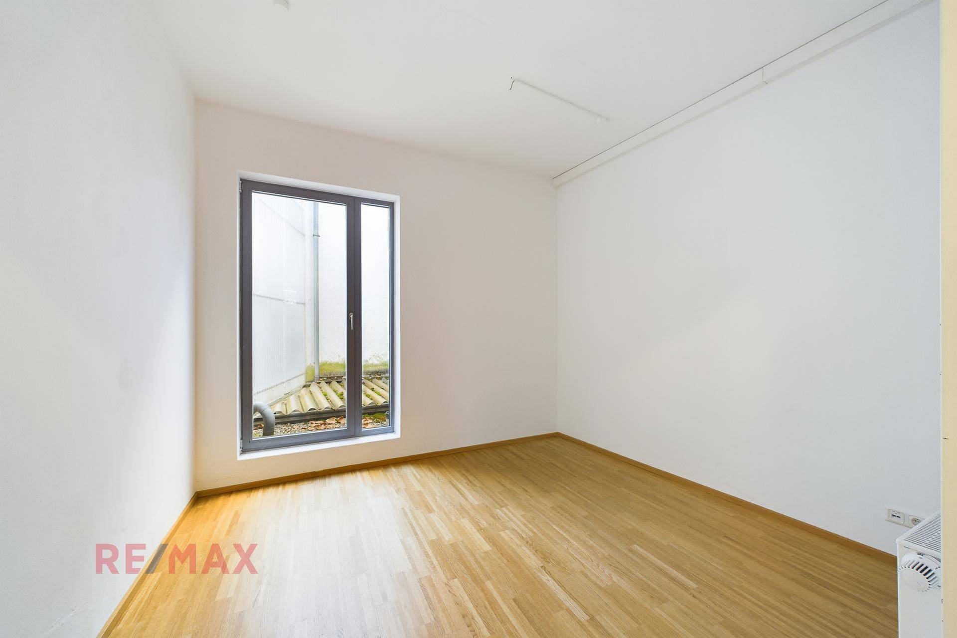 Kleinerer Büroraum mit einem großen Fenster, das Tageslicht spendet, und Holzboden.