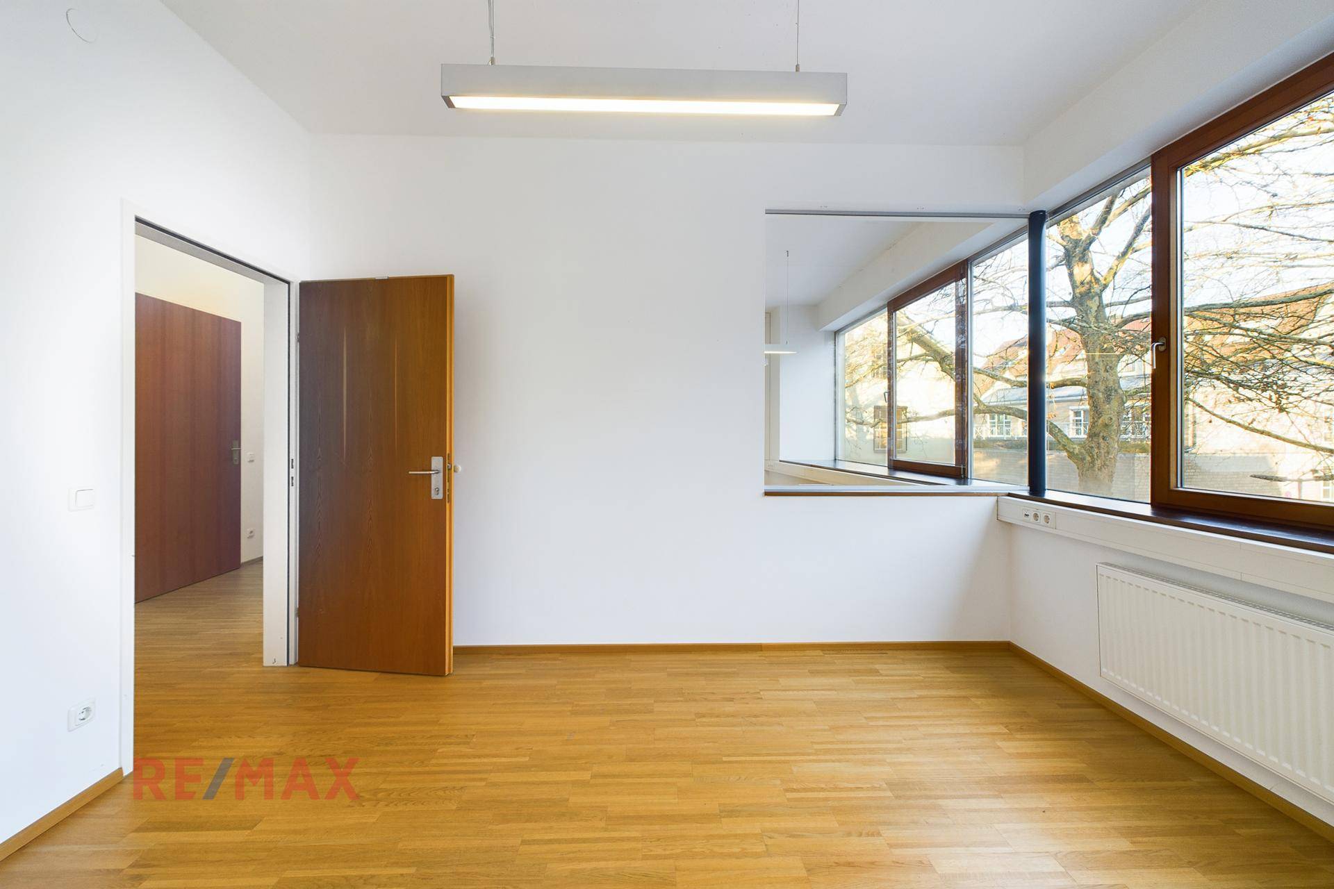 Büroraum mit Holzboden, großen Fenstern und einer Holztür, die zum Flur führt.