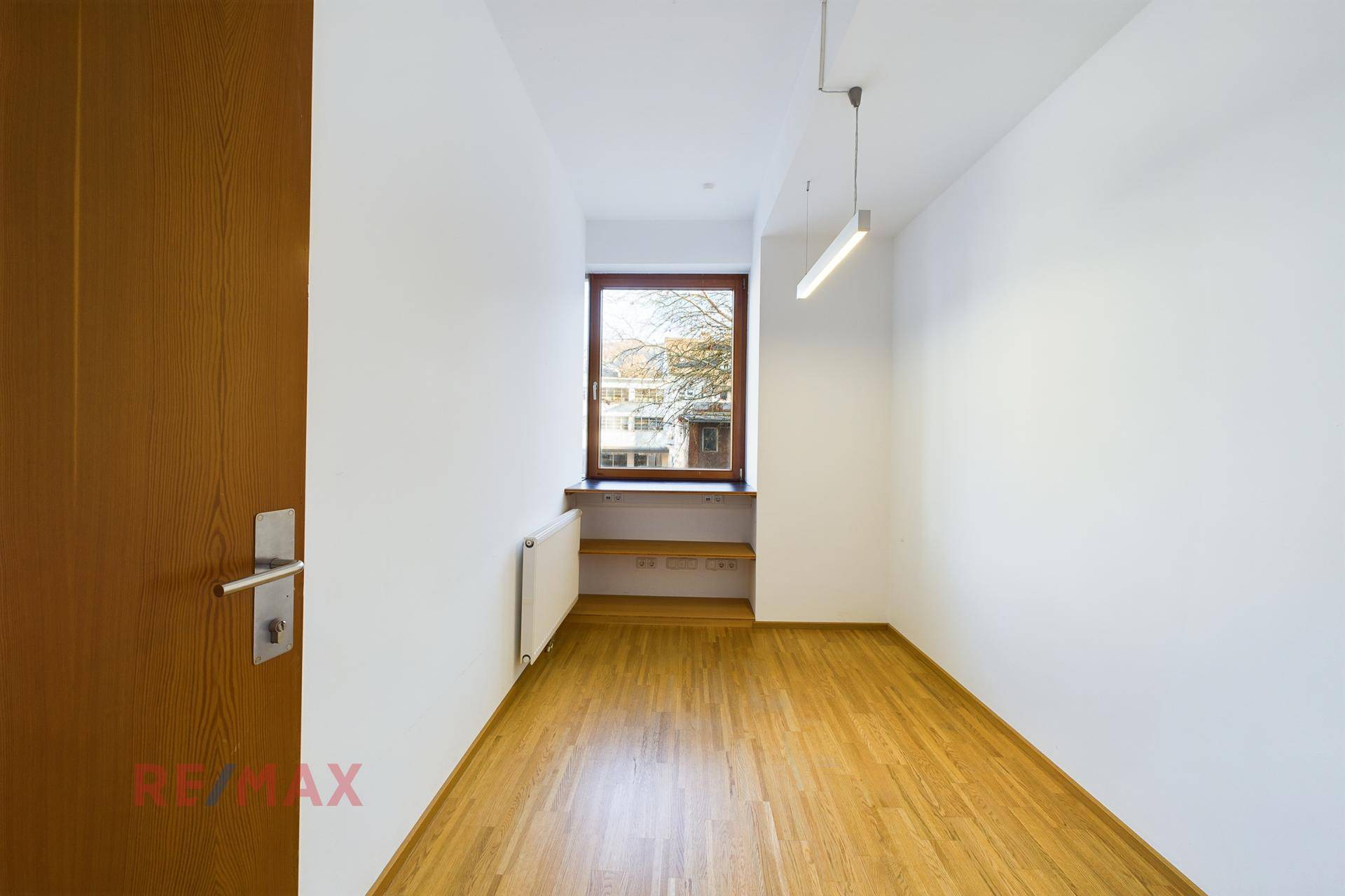 Kompakter Büroraum mit Holzboden, einem Fenster und einer Heizung unter der Fensterbank.