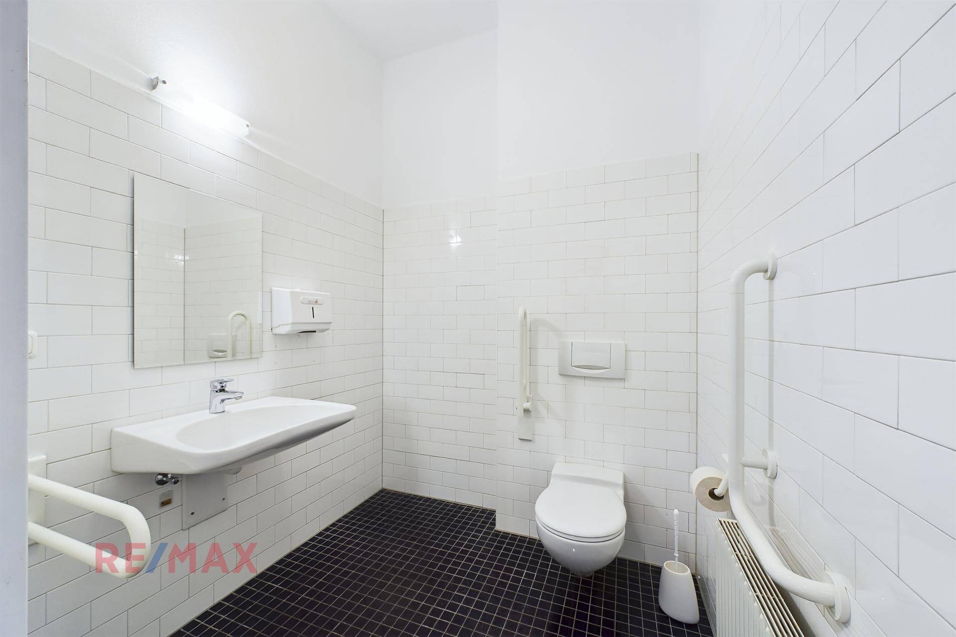 Barrierefreies Badezimmer mit weißen Fliesen, schwarzem Boden und Haltegriffen an der Toilette und am Waschbecken.