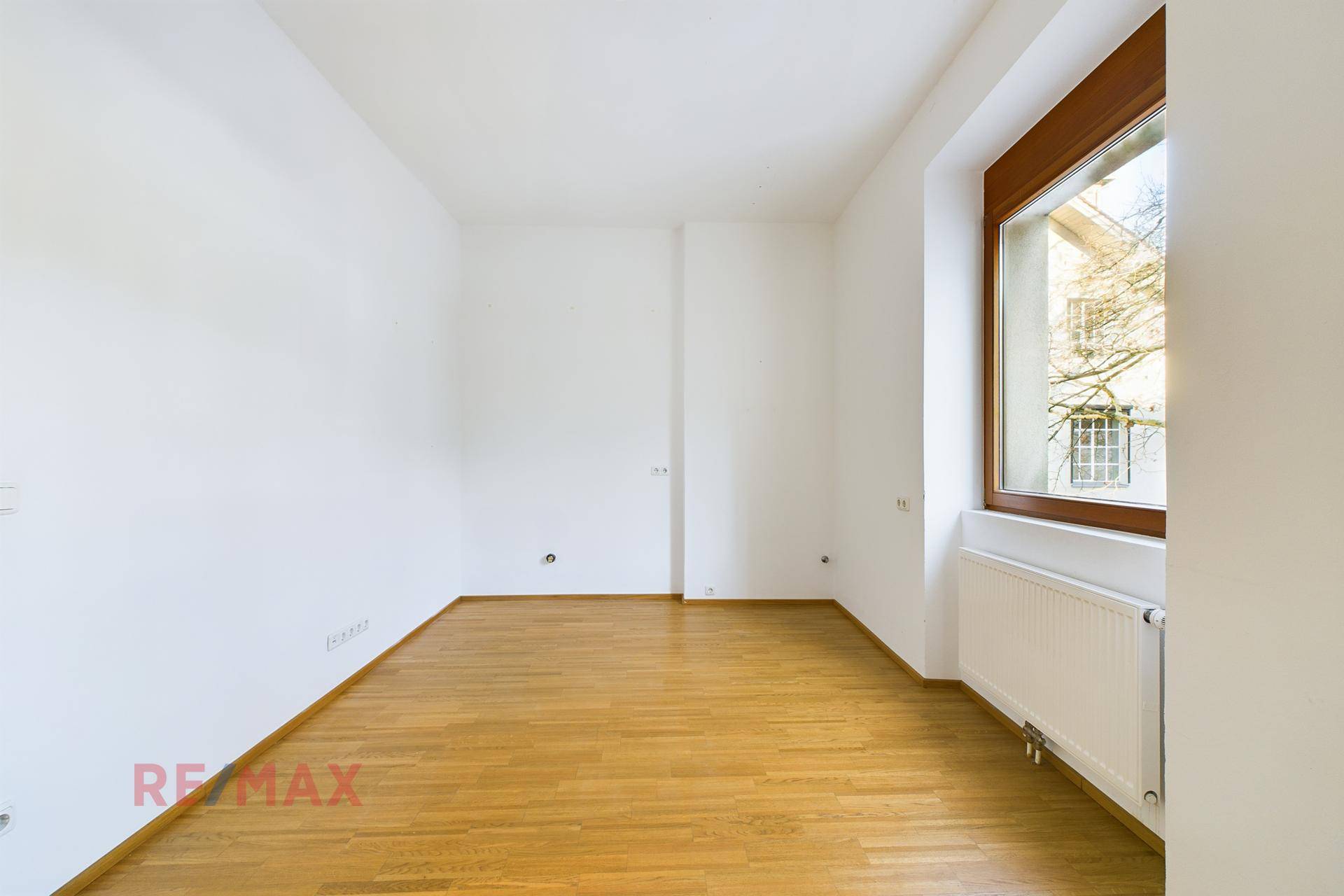Leerer Büroraum mit Holzboden, weißen Wänden und einem Fenster mit Blick nach draußen.