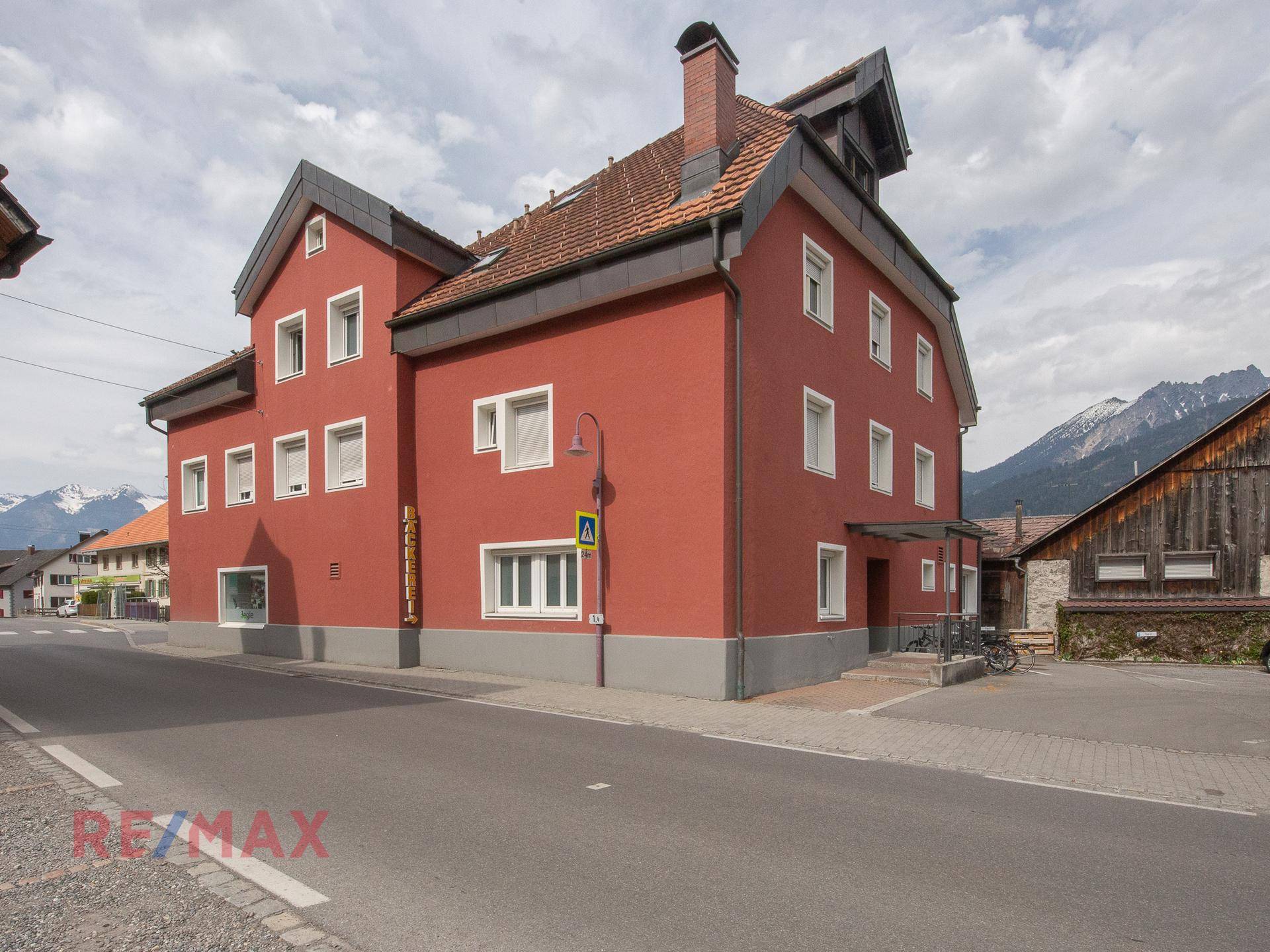 Rotes Gebäude mit traditionellem Dach und Fenstern, umgeben von ländlicher Architektur und Bergen.