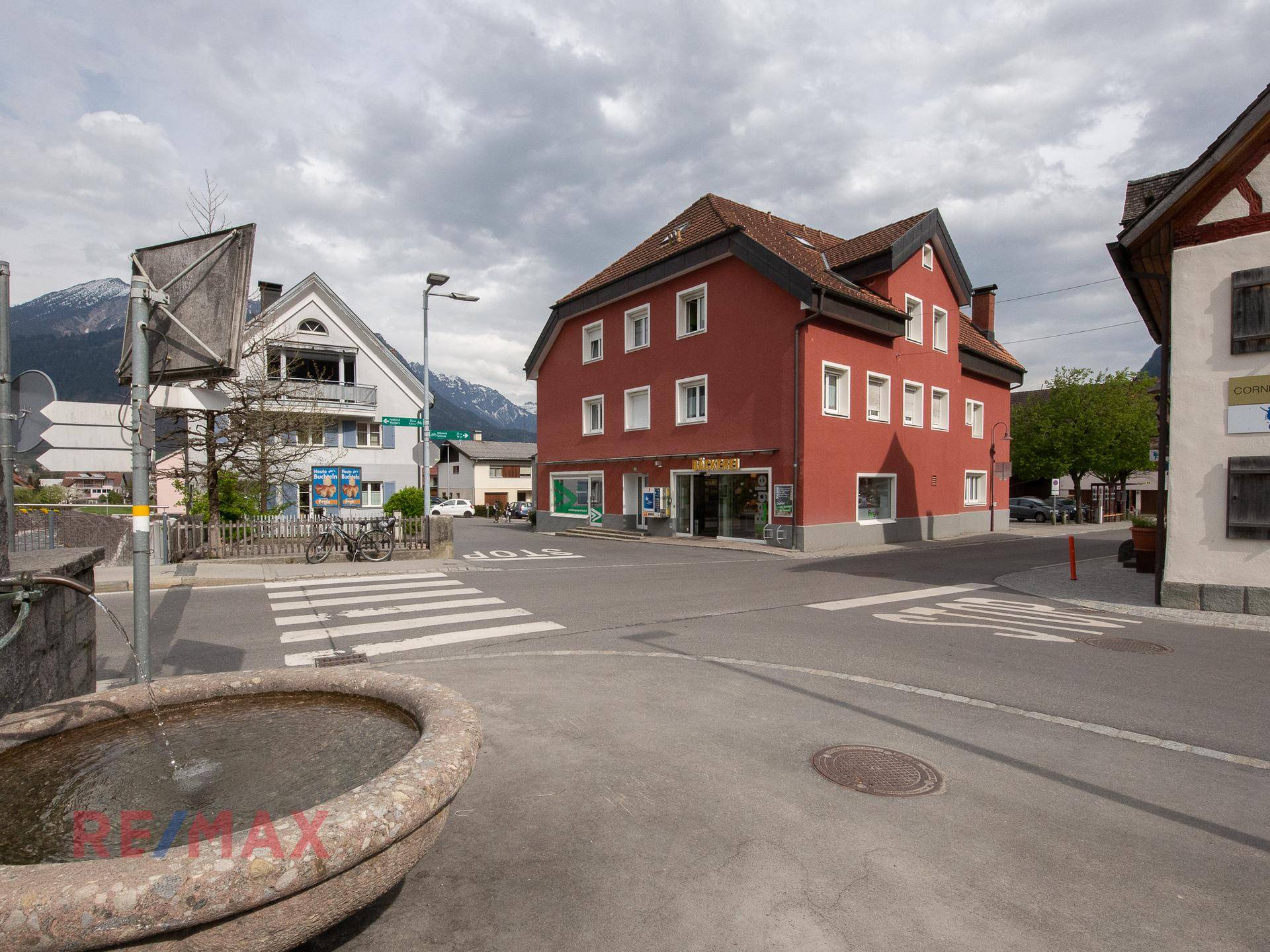 Rotes Gebäude an einer ländlichen Kreuzung mit einem Brunnen im Vordergrund und Bergblick.