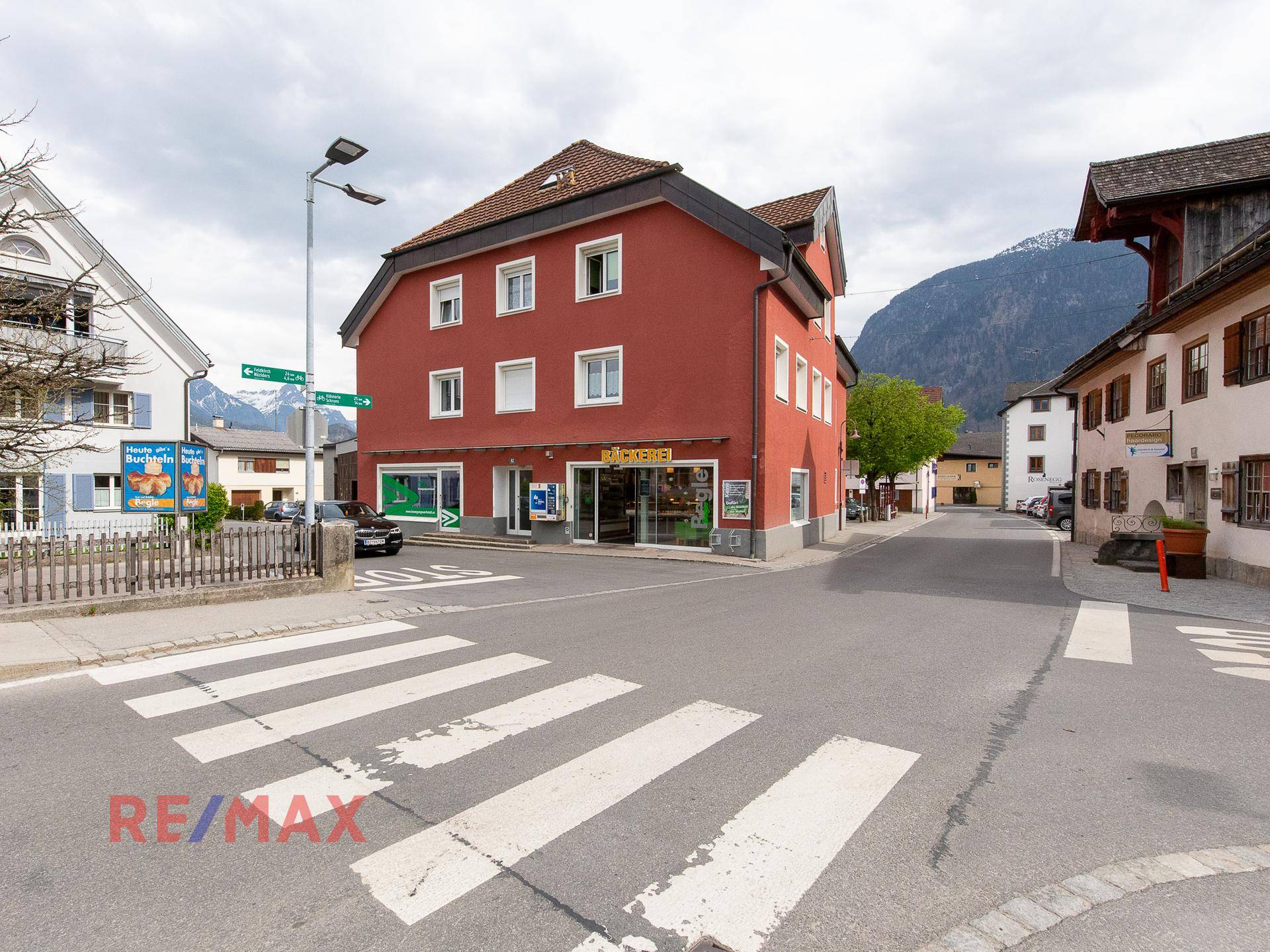 Rotes Gebäude an einer Straßenkreuzung mit Zebrastreifen und umliegender ländlicher Bebauung.