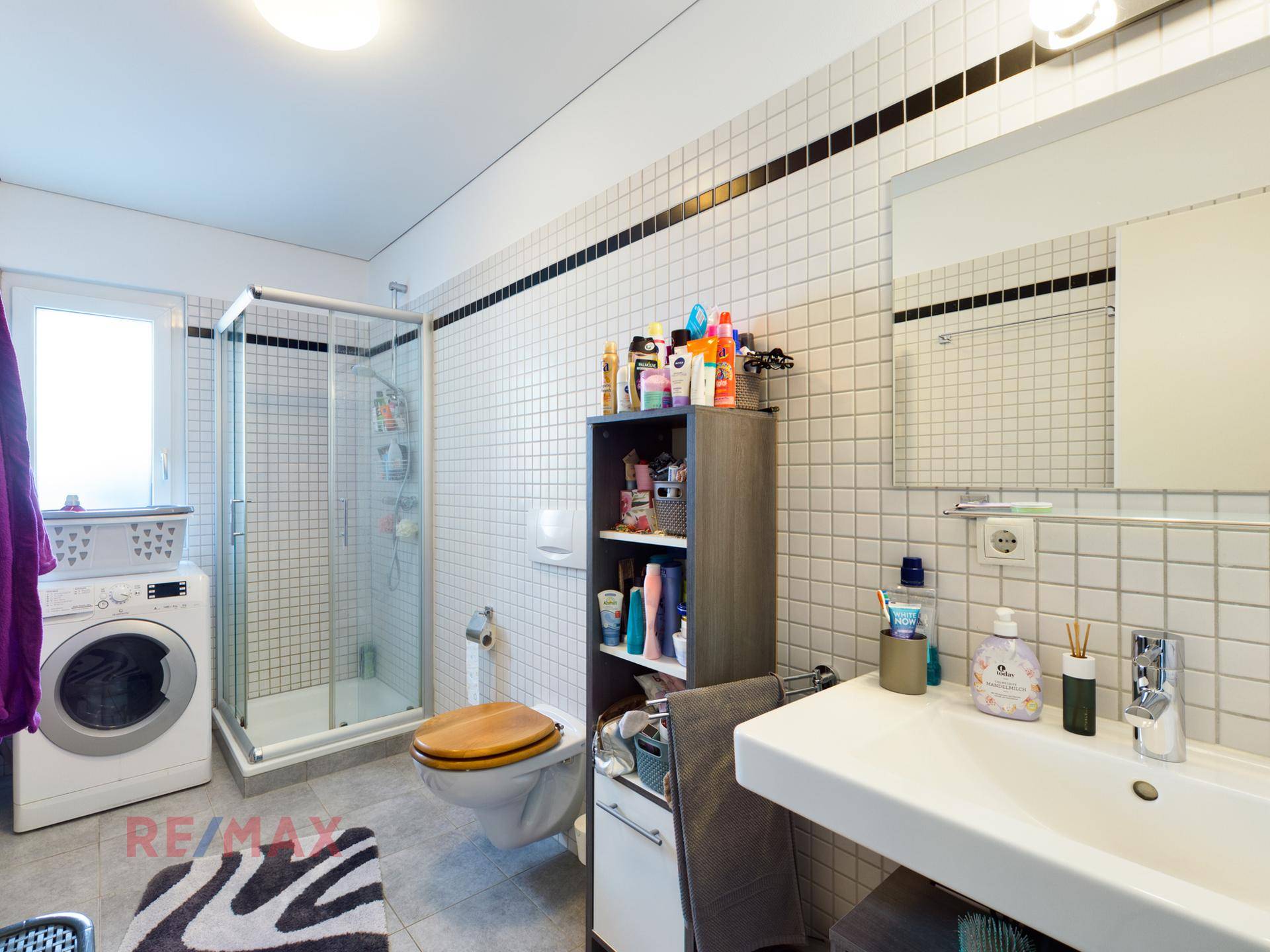 Badezimmer mit Duschkabine, Waschbecken, WC und Waschmaschine, gefliest in Weiß.