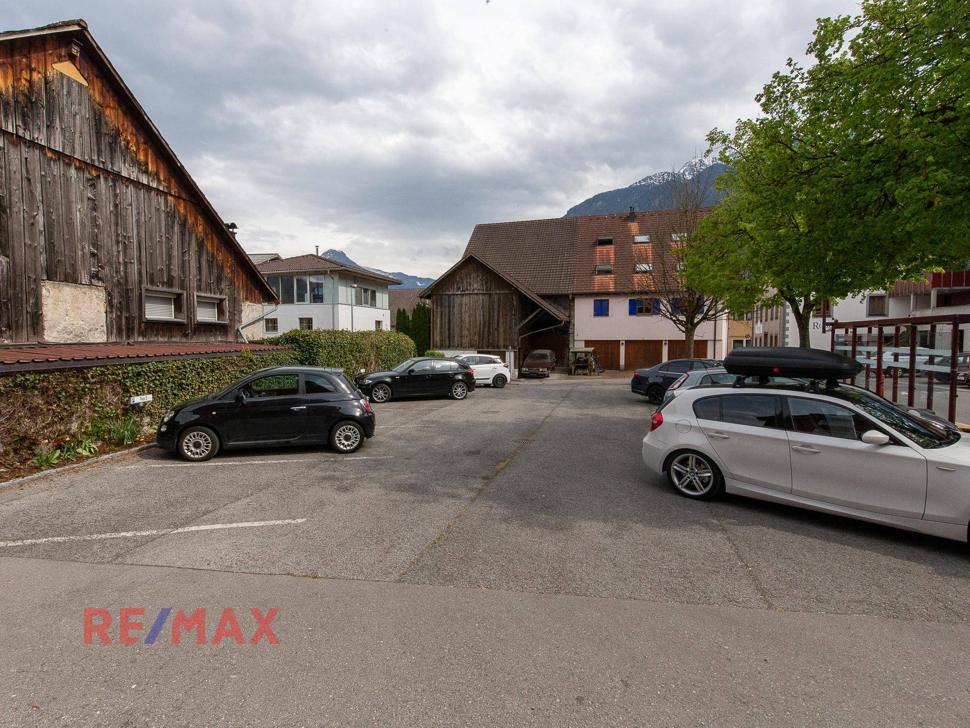 Parkplatz mit mehreren Autos, umgeben von traditionellen Gebäuden und grüner Landschaft.