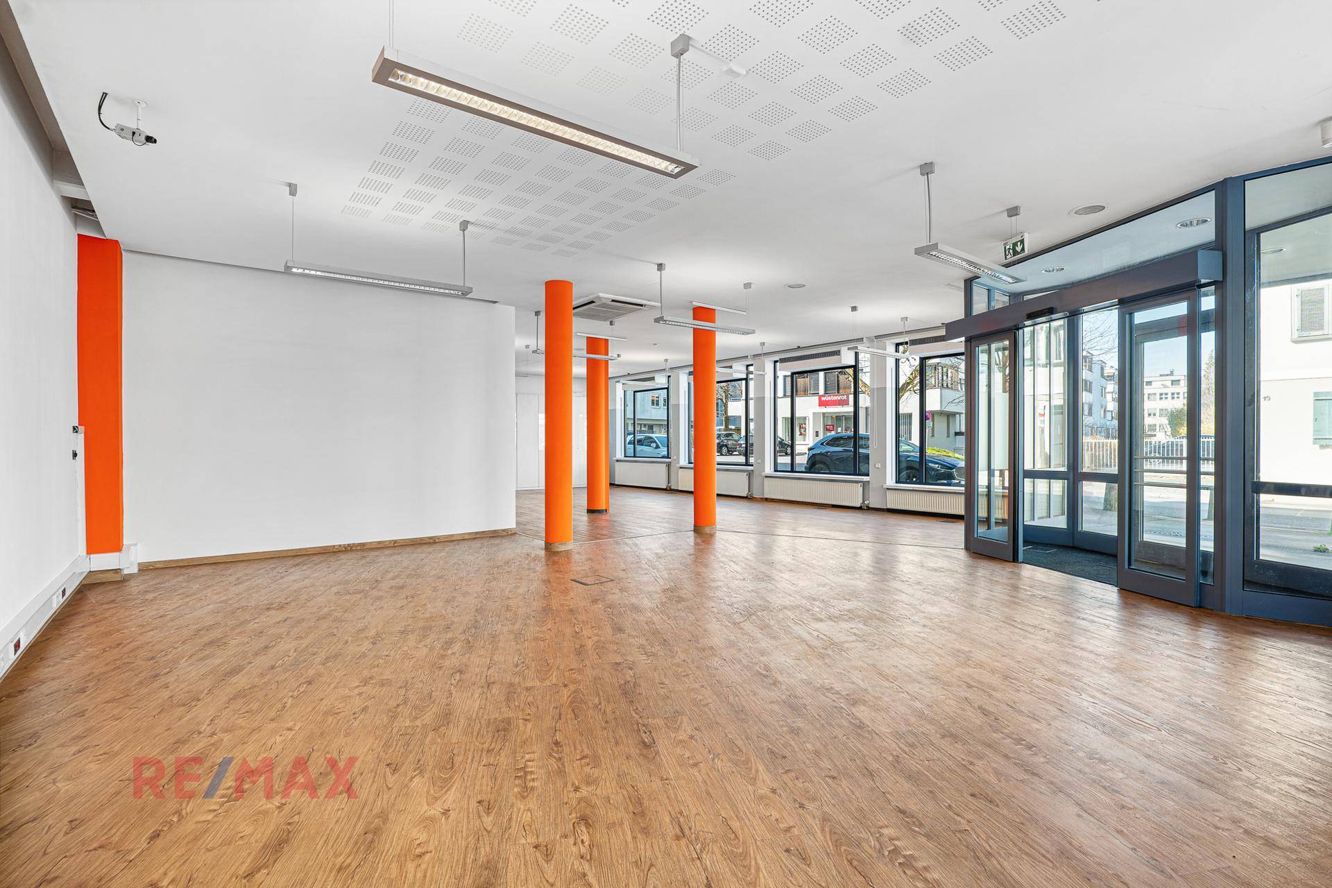 Weitläufiger Geschäftsraum mit vielen Fenstern, modernen Deckenleuchten und orangefarbenen Akzentsäulen.
