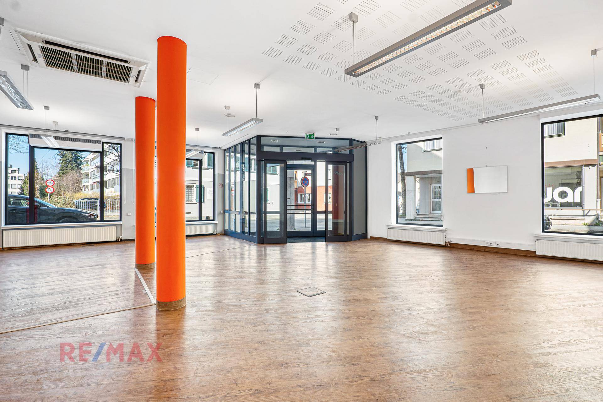 Geräumiger, leerer Geschäftsraum mit großen Fenstern, orangefarbenen Säulen und hellem Holzboden.
