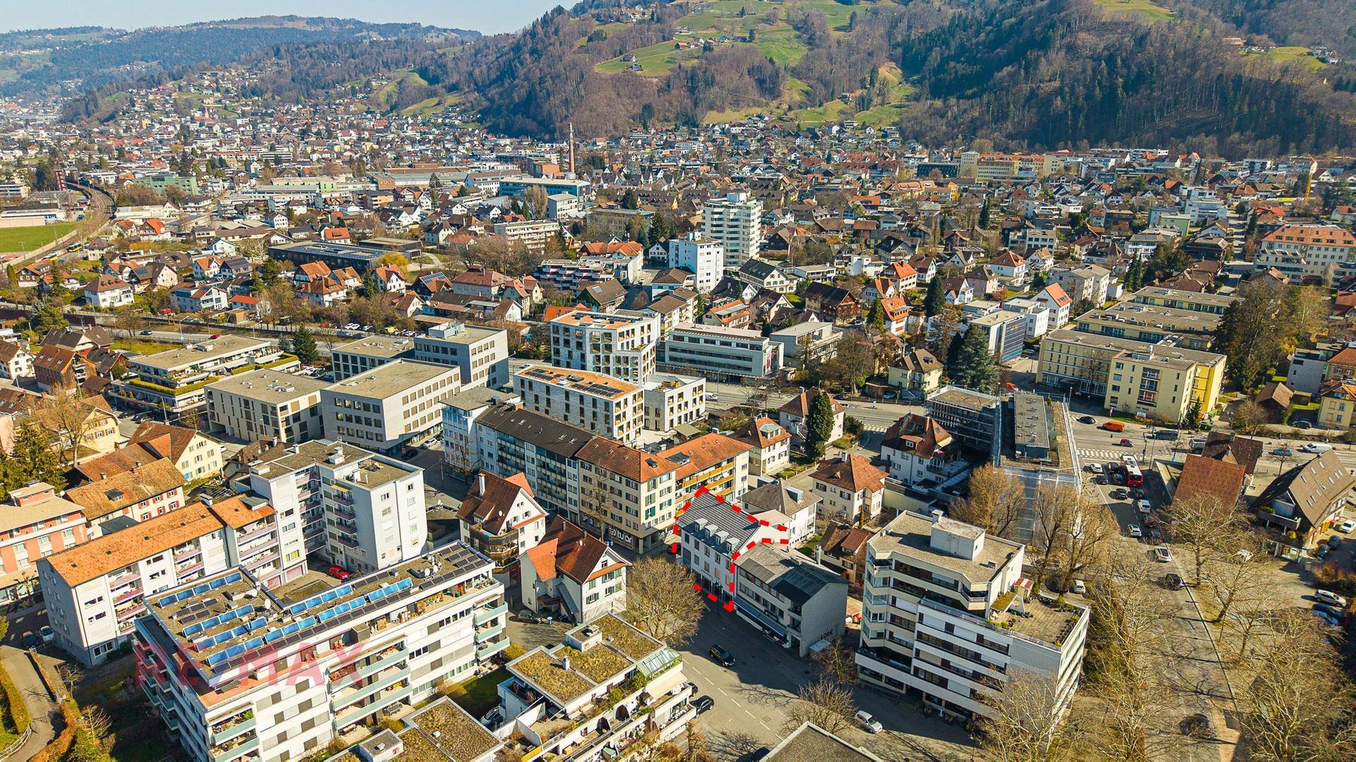 Weitläufige Luftaufnahme der Stadt mit dem umrandeten Gebäude, umgeben von Wohn- und Geschäftsgebäuden sowie Bergen.