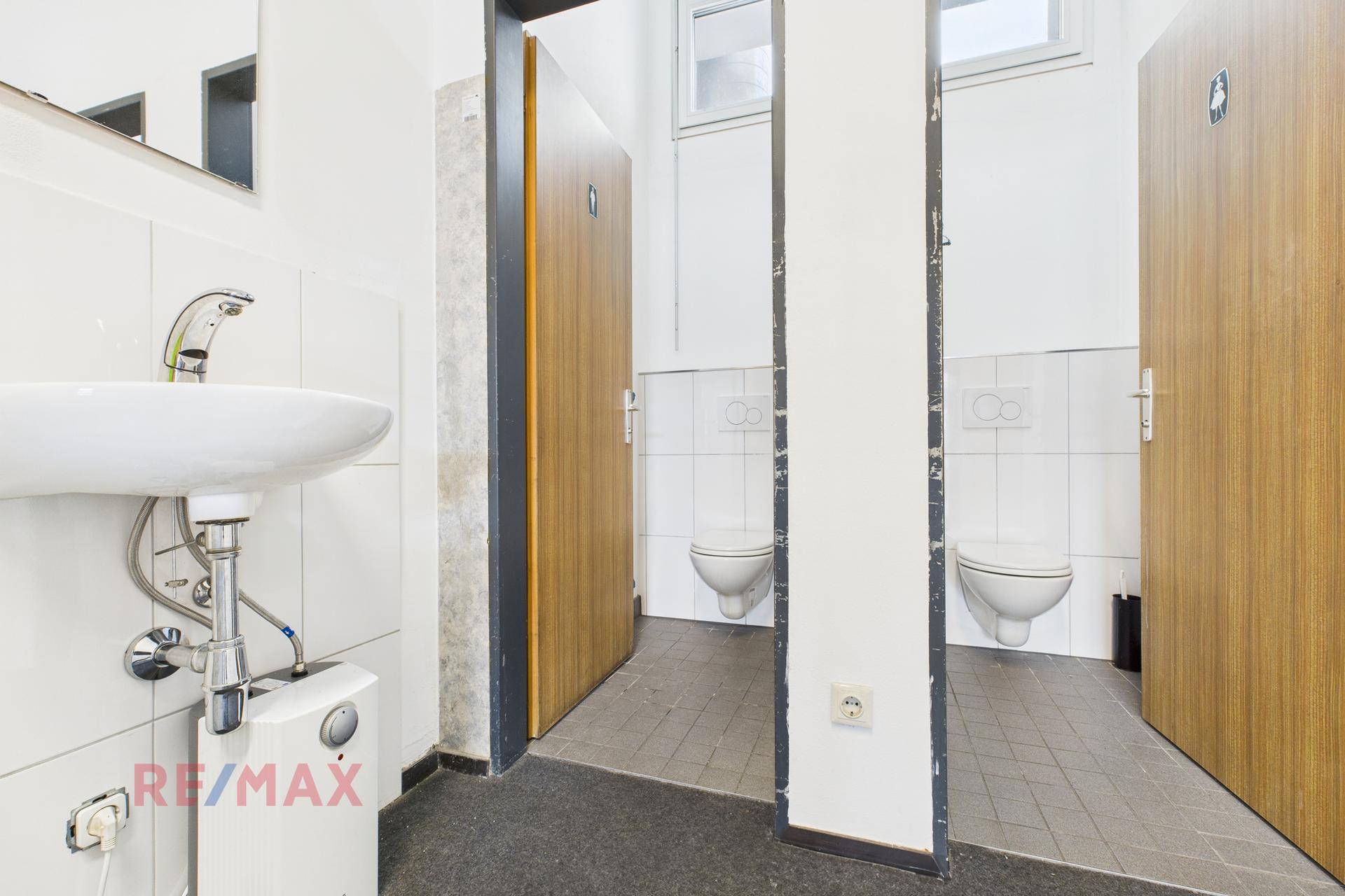 Waschbereich mit Waschbecken und Zugang zu zwei Toilettenkabinen mit weißen Fliesen.