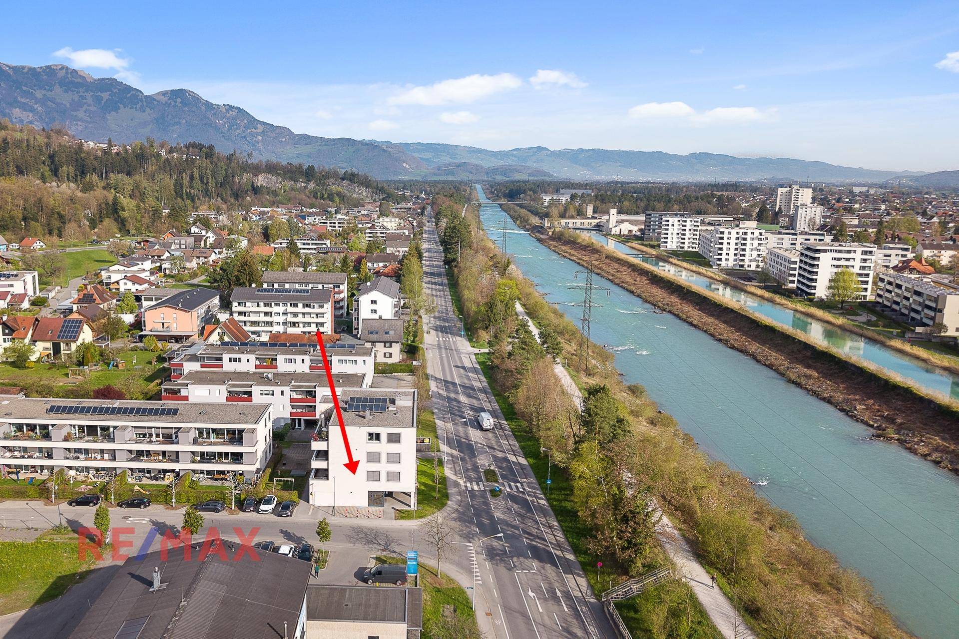 Luftaufnahme des Geschäftsgebäudes in Flussnähe mit umliegender Bebauung und Bergkulisse.