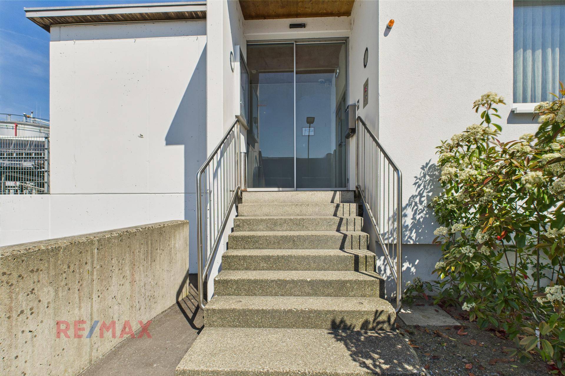 Eingangsbereich eines modernen Gebäudes mit Steintreppe, Edelstahlgeländer und einer großen Glastür.