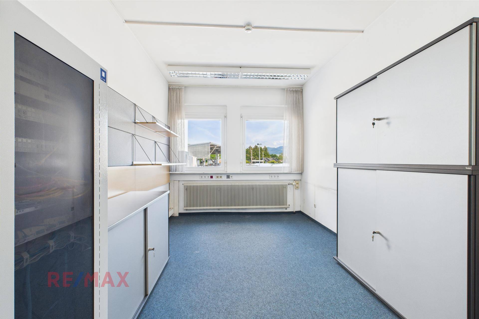 Einzelbüro mit blauem Teppichboden, großen Fenstern und praktischem Stauraum, bietet einen angenehmen Blick ins Grüne.