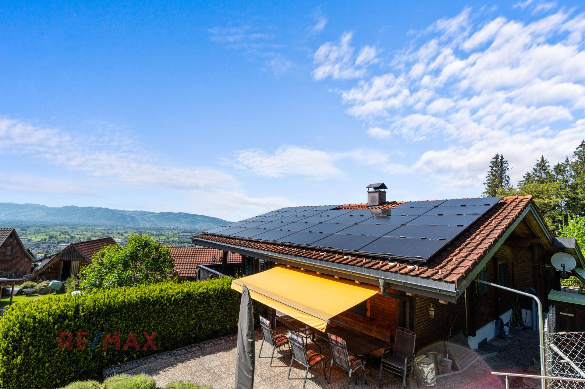 Haus mit Solaranlage auf dem Dach und einer kleinen Terrasse mit Markise und Sitzgelegenheiten.