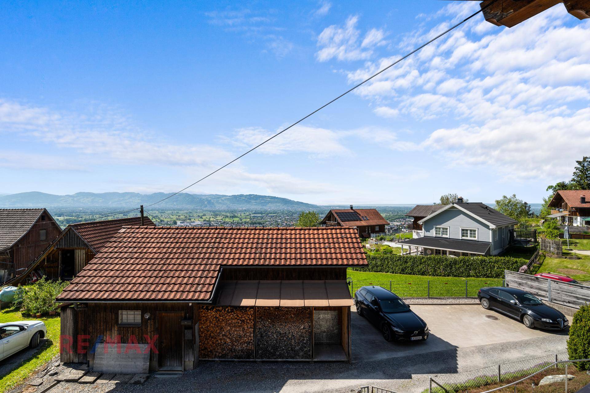 Rustikales Holzhaus mit Ziegeldach, Holzstapel und geparkten Autos in ländlicher Umgebung.