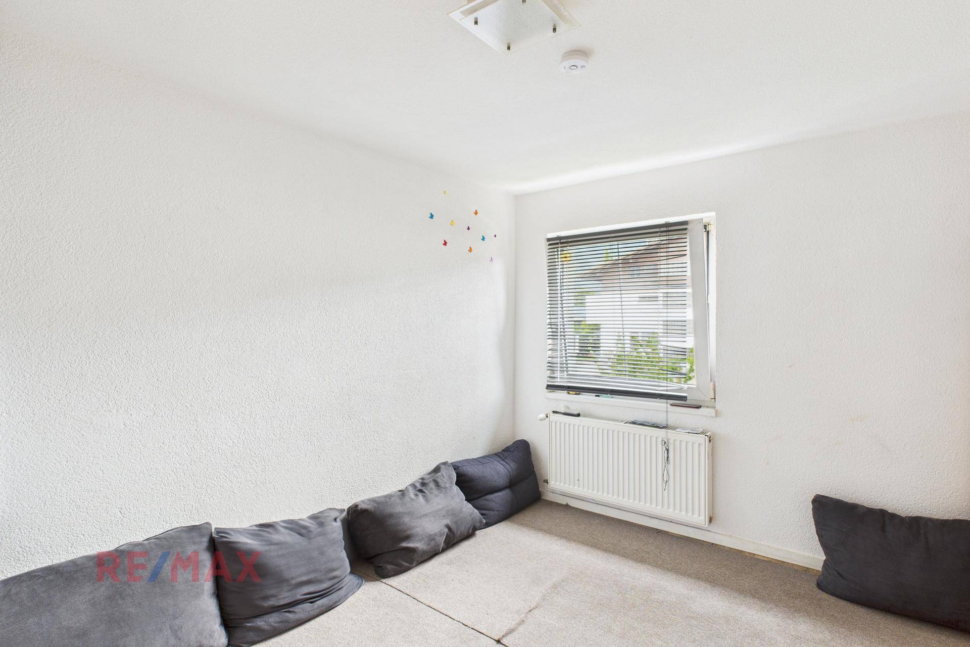 Kleines Zimmer mit Teppichboden, einem Fenster mit Jalousien und grauen Bodenkissen.