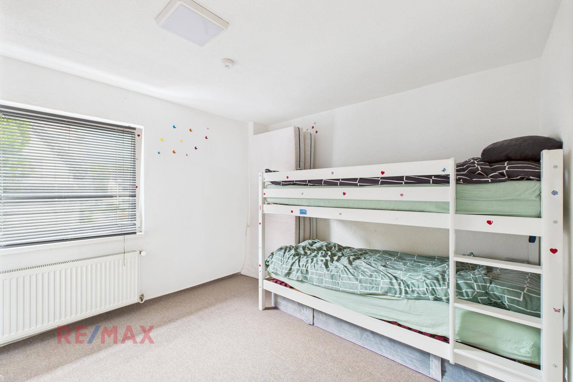 Kinderzimmer mit Etagenbett, Teppichboden und einem Fenster mit Jalousien.