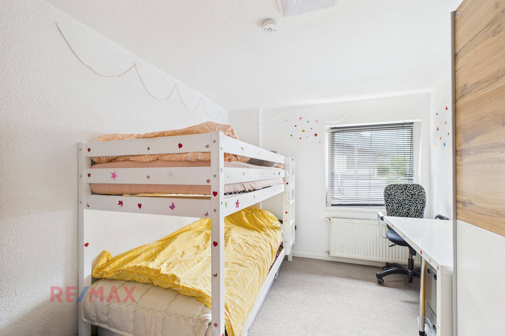 Helles Kinderzimmer mit Etagenbett, Schreibtisch und einem Fenster mit Jalousien.