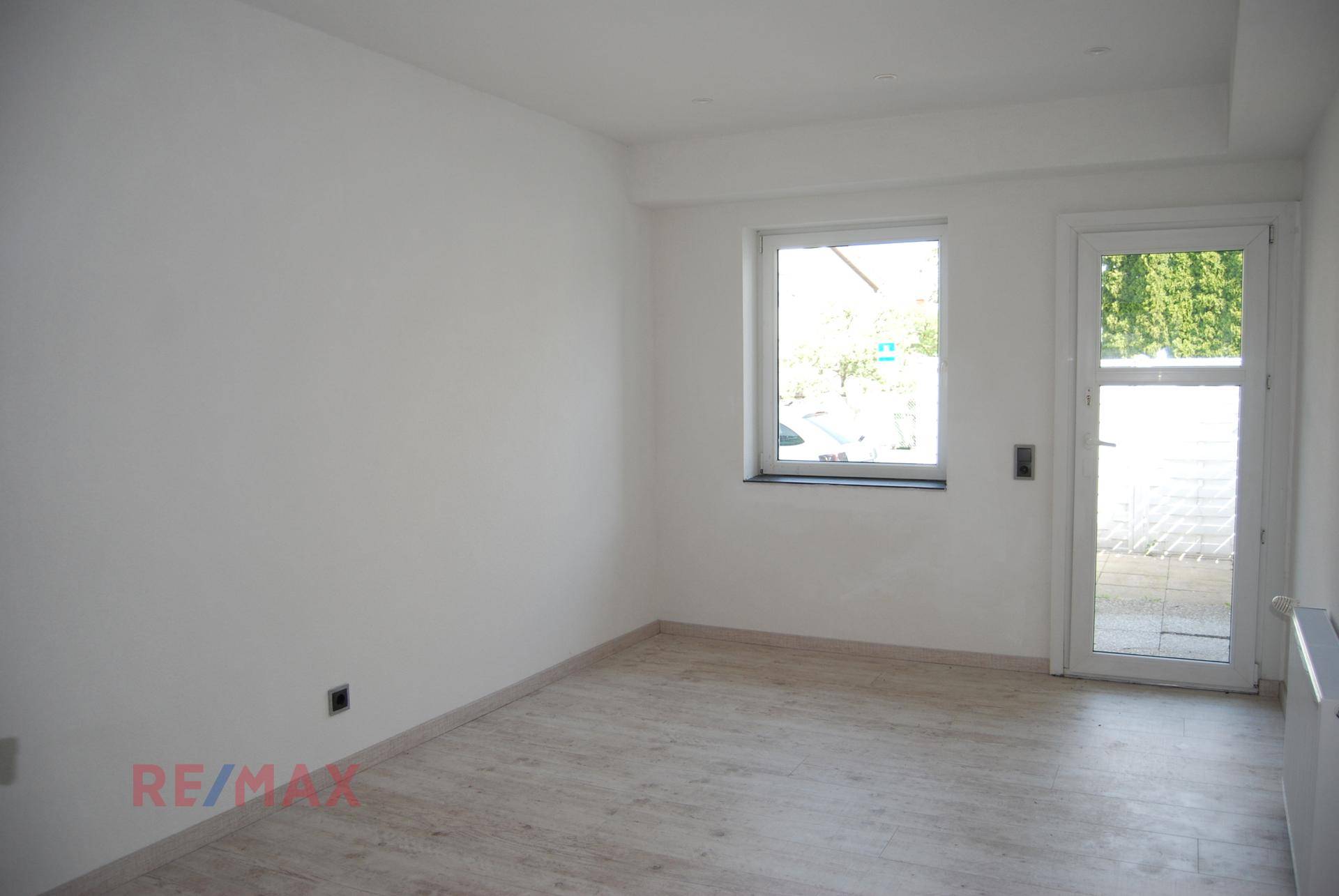 Leerer Raum mit hellem Laminatboden, weiß gestrichenen Wänden und einem Fenster zur Außenseite.