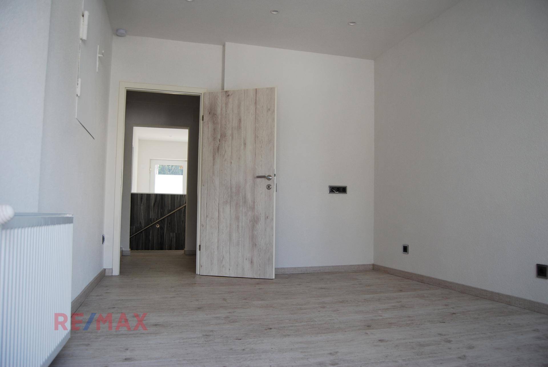 Schlichter Raum mit hellem Laminatboden, weißen Wänden und einer Holztür zu einem weiteren Bereich.