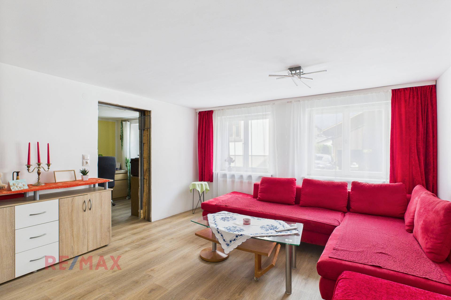 Helles Wohnzimmer mit rotem Sofa, großen Fenstern und Zugang zu einem weiteren Raum.
