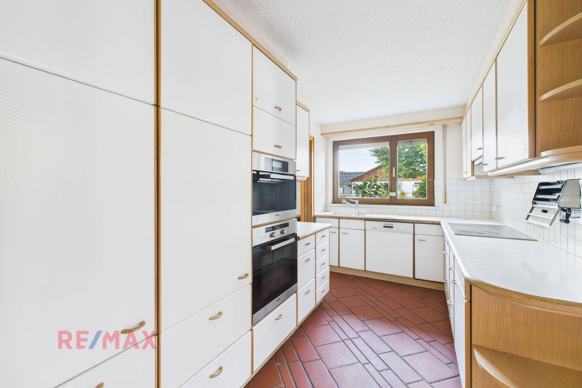 Helle Küche mit weißen Einbauschränken, Fenster und Terrakottafliesen, gut beleuchtet.