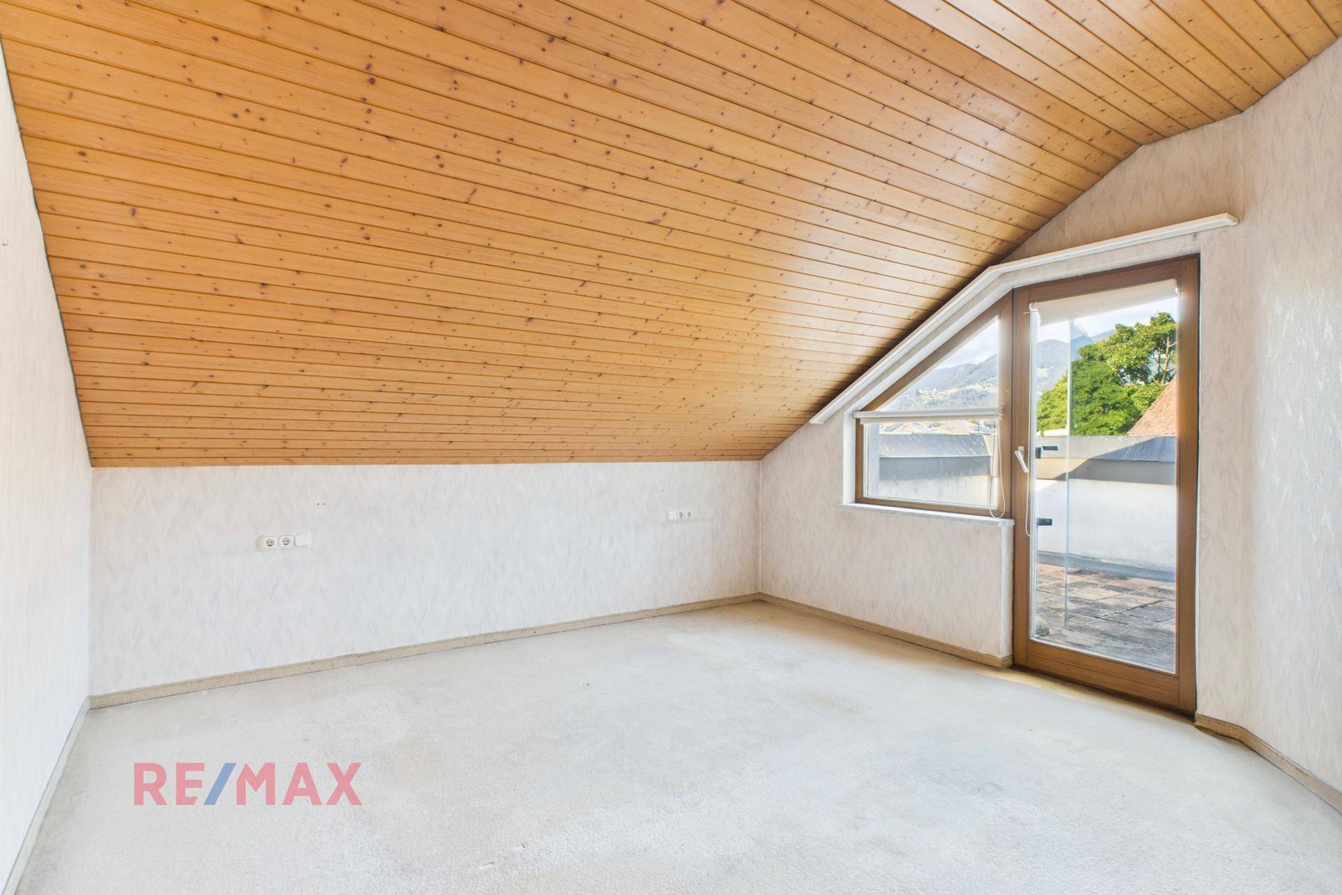 Dachzimmer mit Holzdecke, Teppichboden und Bergblick durch das Fenster.