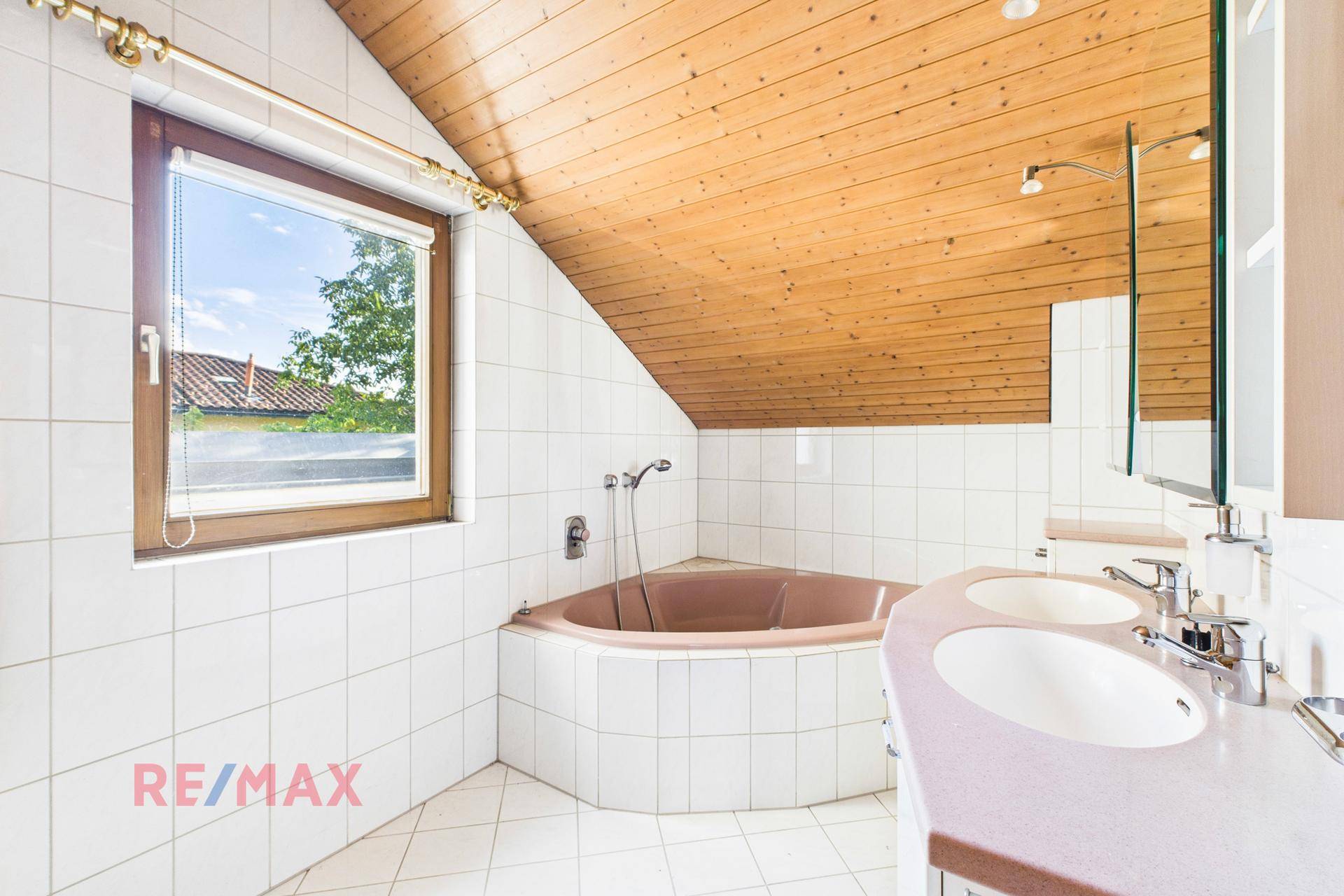 Badezimmer mit Eckbadewanne, Doppelwaschbecken und Holzdecke, hell gefliest.