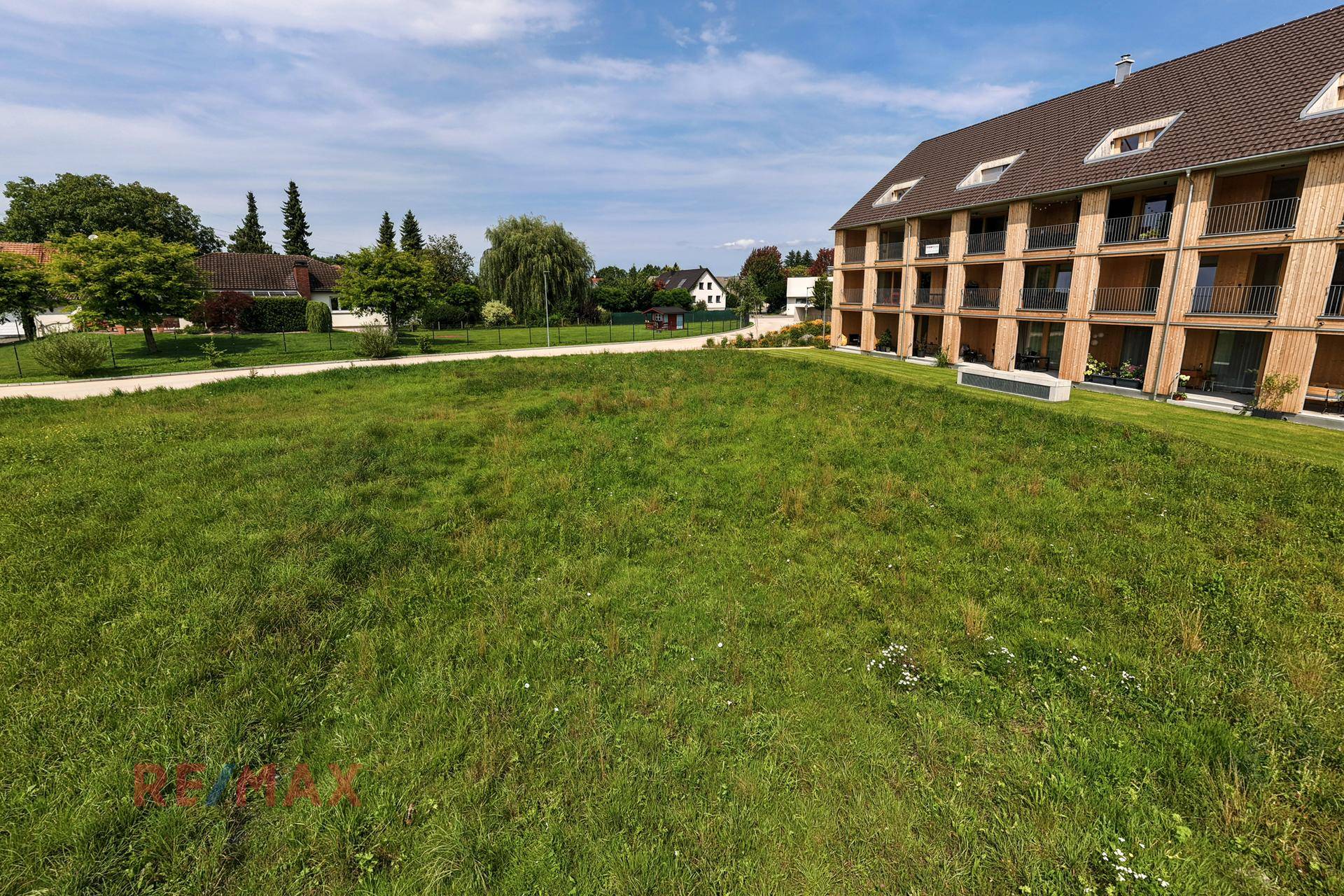 Grüne Wiese als Baugrundstück neben einem modernen Mehrfamilienhaus unter blauem Himmel.