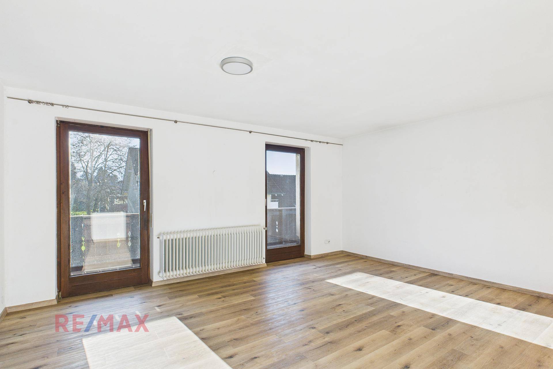Helles Wohnzimmer mit Parkettboden und zwei Fenstertüren, die auf einen Balkon führen.