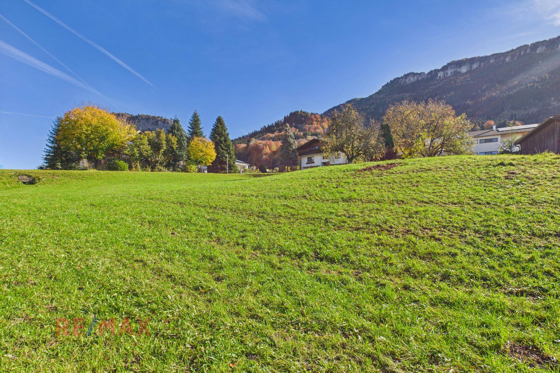 Sonnige Wiesenlandschaft mit Bäumen und Häusern im Hintergrund, umgeben von Bergen.