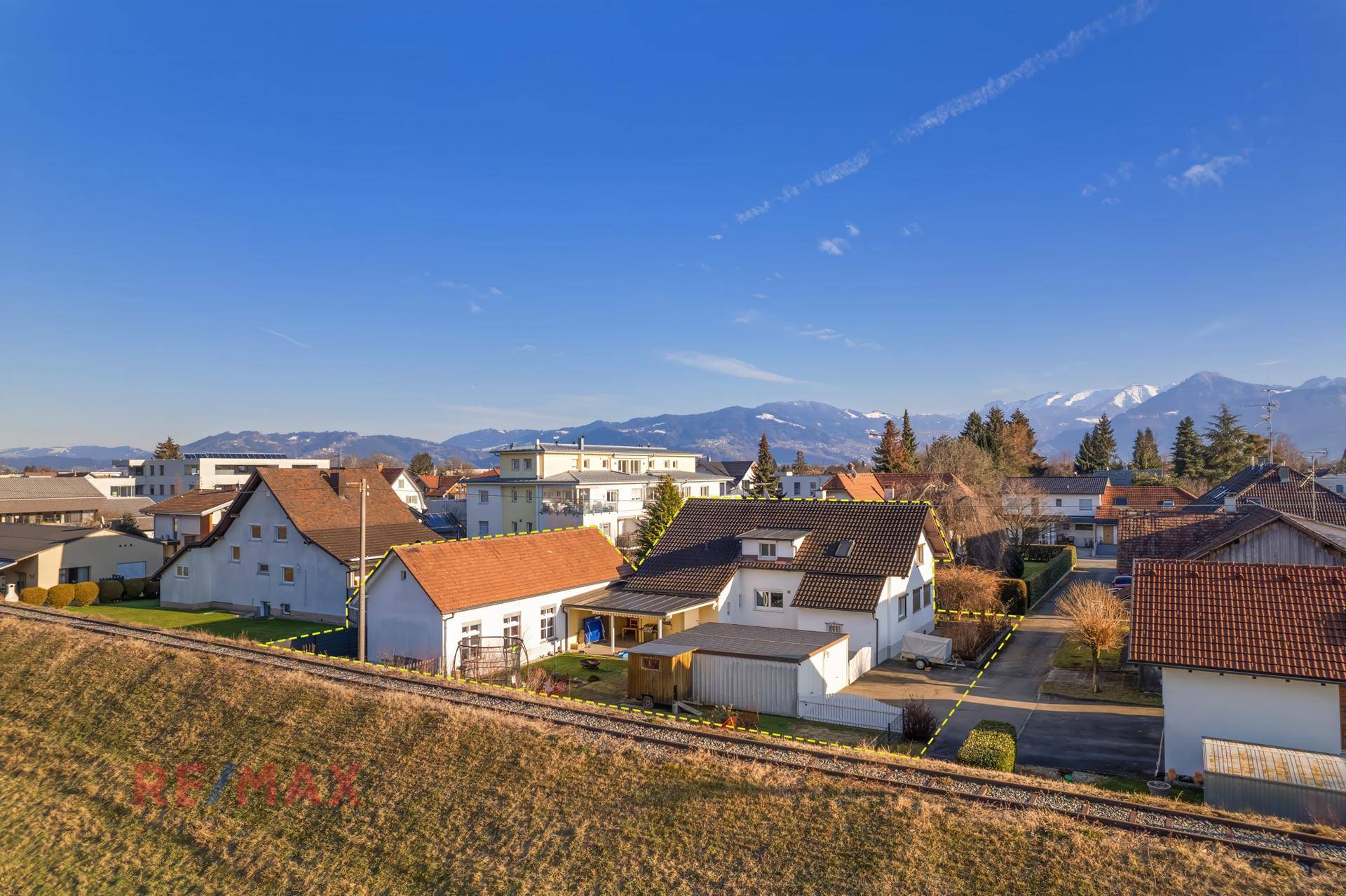 Luftaufnahme des Einfamilienhauses mit umliegenden Gebäuden und Bergen im Hintergrund.