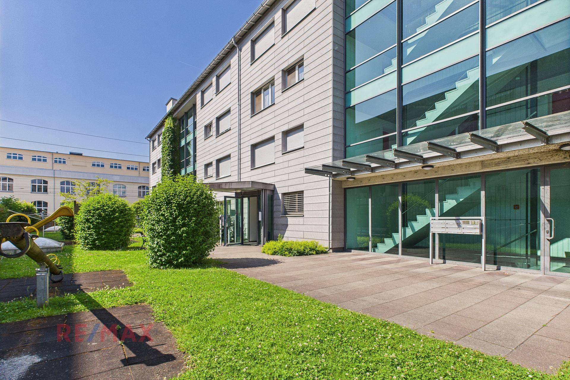 Modernes Gebäude mit Glasfassade und begrüntem Außenbereich unter blauem Himmel.