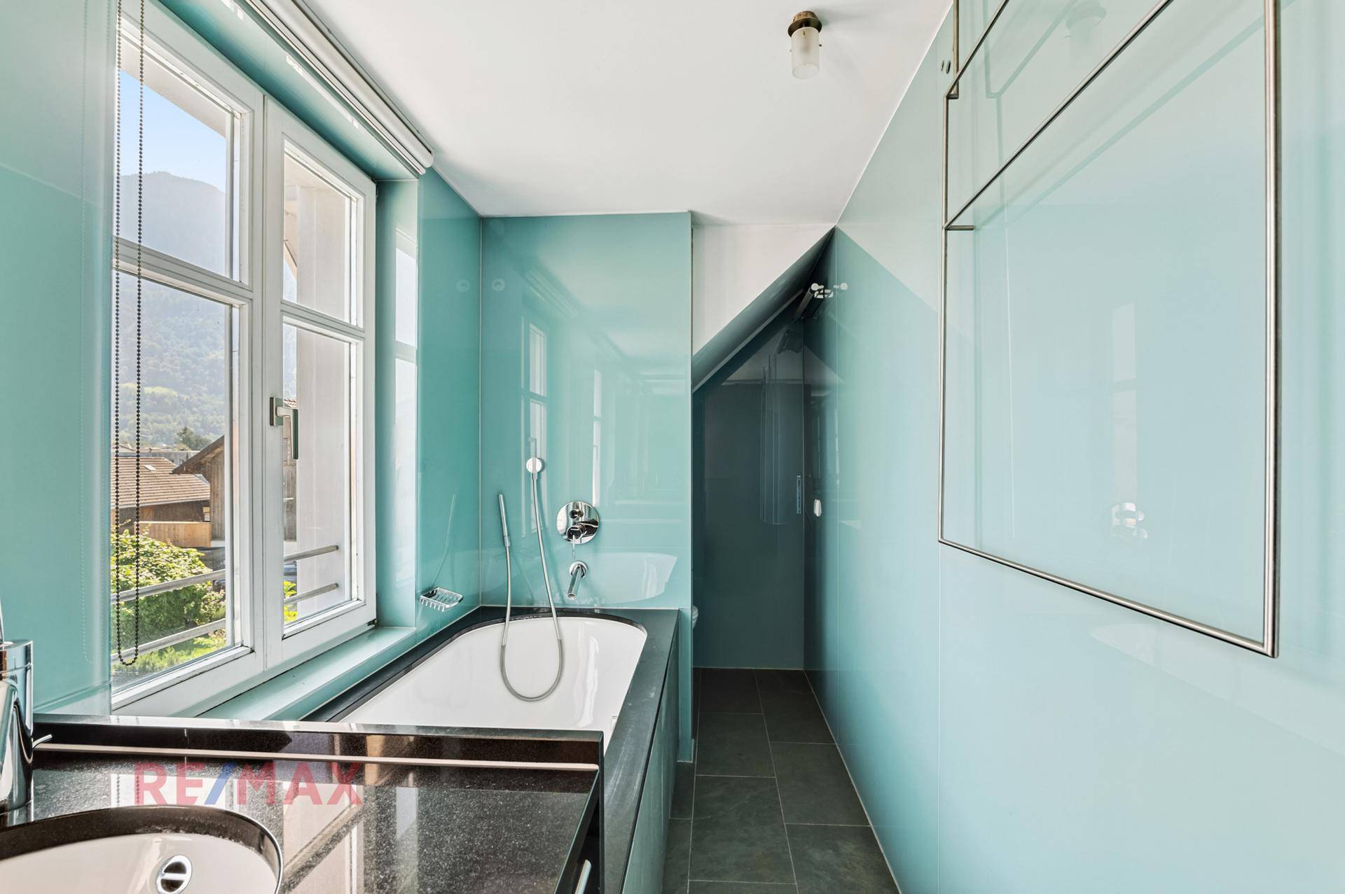 Modernes Badezimmer mit blauen Glaswänden, Badewanne, Dusche und Fenster mit Bergblick.