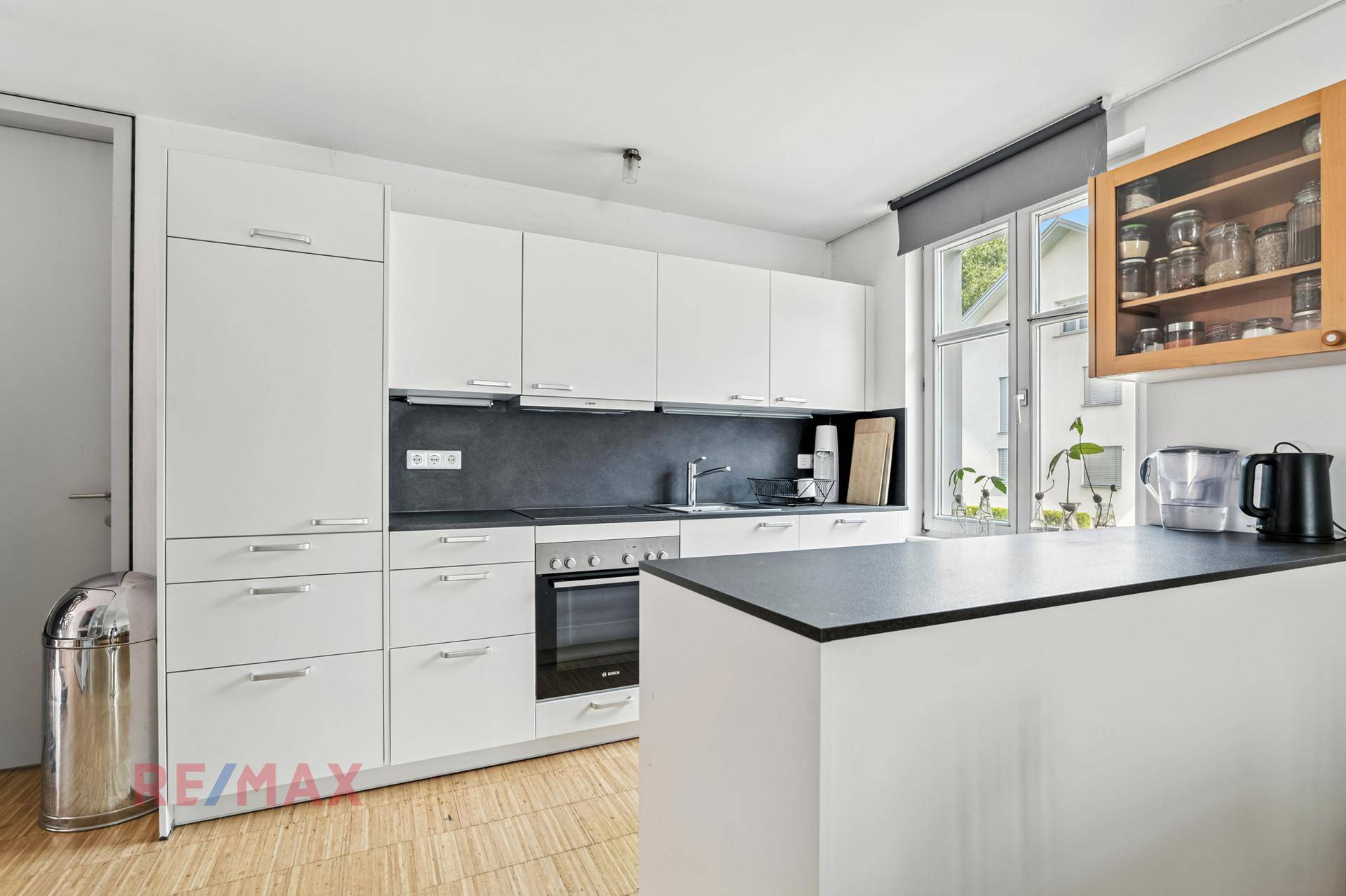 Funktionale weiße Küche mit modernen Geräten, dunkler Arbeitsplatte und Fenster.