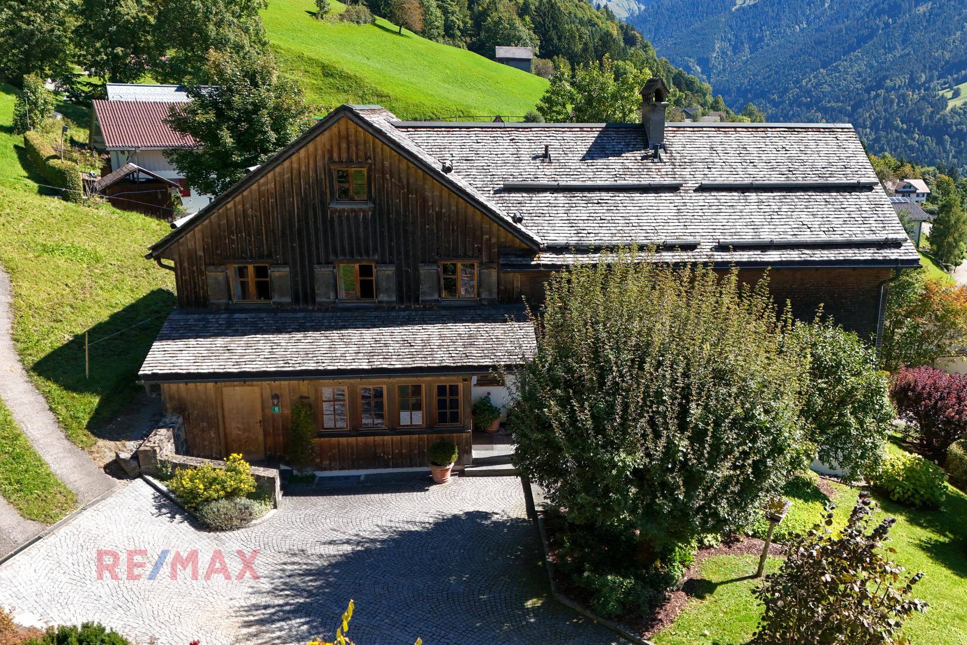 Traditionelles Holzhaus mit Schindeldach, umgeben von grünen Hügeln und Bäumen in alpiner Landschaft.