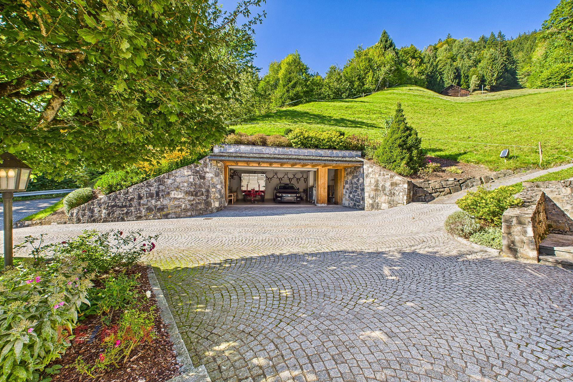 Einfahrt mit Kopfsteinpflaster und integrierter Garage in Hanglage, umgeben von viel Grün.
