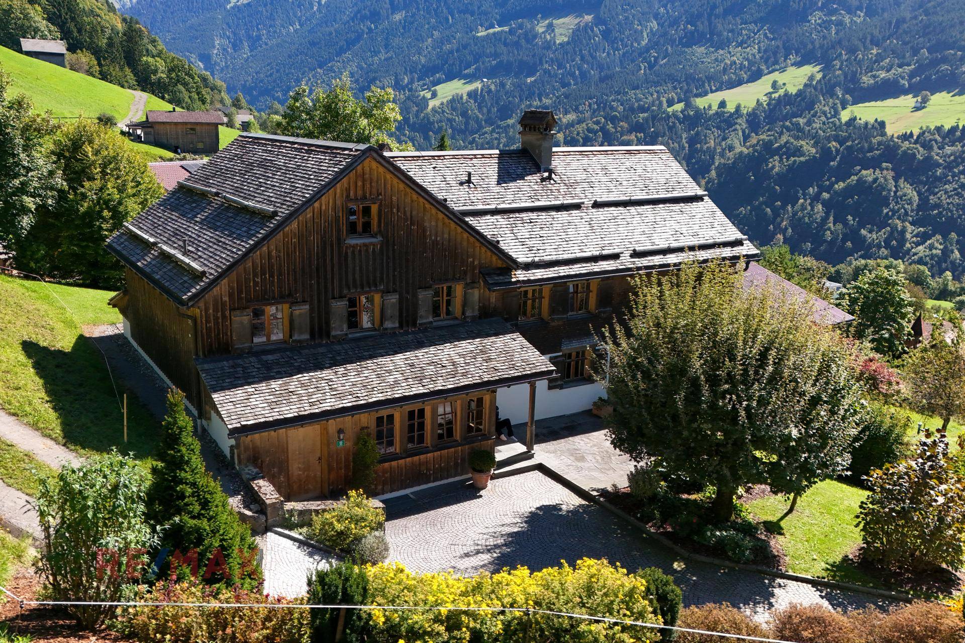 Traditionelles Holzhaus mit Schindeldach, umgeben von einem gepflegten Garten und grünen Hügeln.