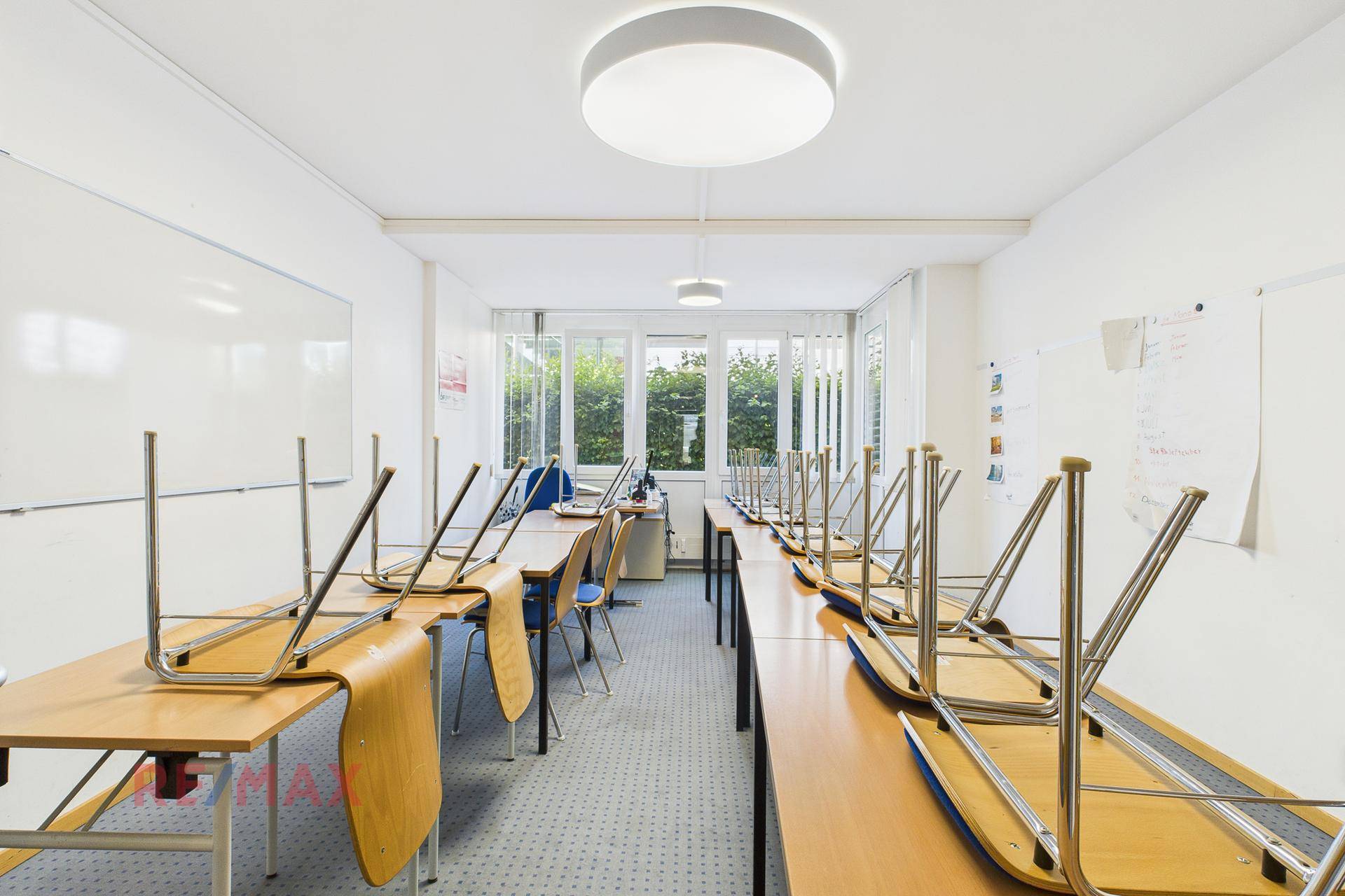 Klassenzimmer mit aufgestellten Tischen und Stühlen, großen Fenstern und hellem Teppichboden.