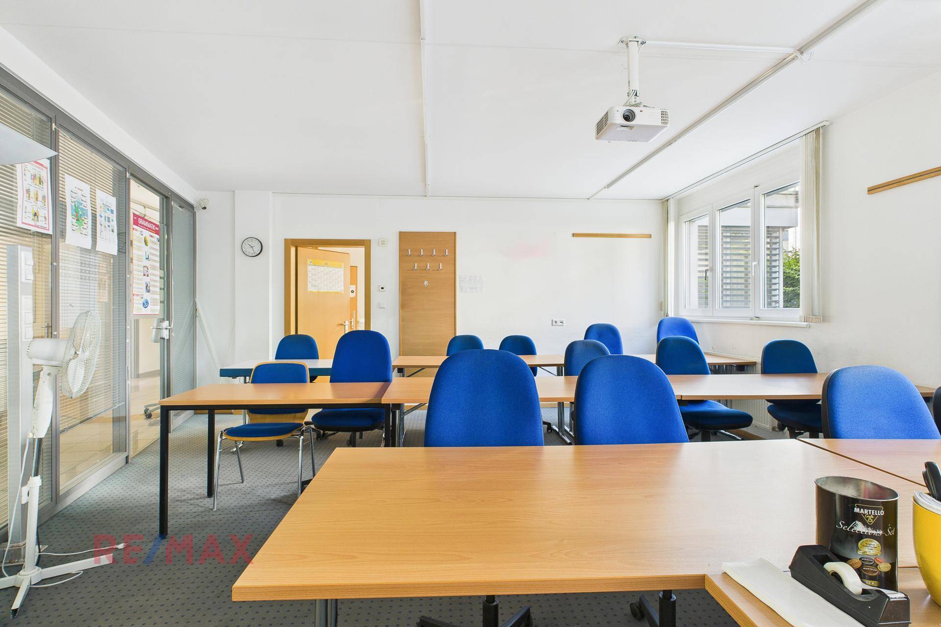 Klassenzimmer mit mehreren Tischen und blauen Stühlen, ausgestattet mit einem Beamer und großen Fenstern.