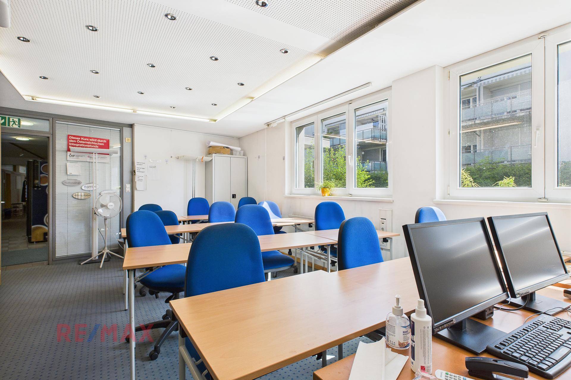 Klassenzimmer mit Tischen, blauen Stühlen und Computermonitoren, bietet viel Tageslicht durch große Fenster.