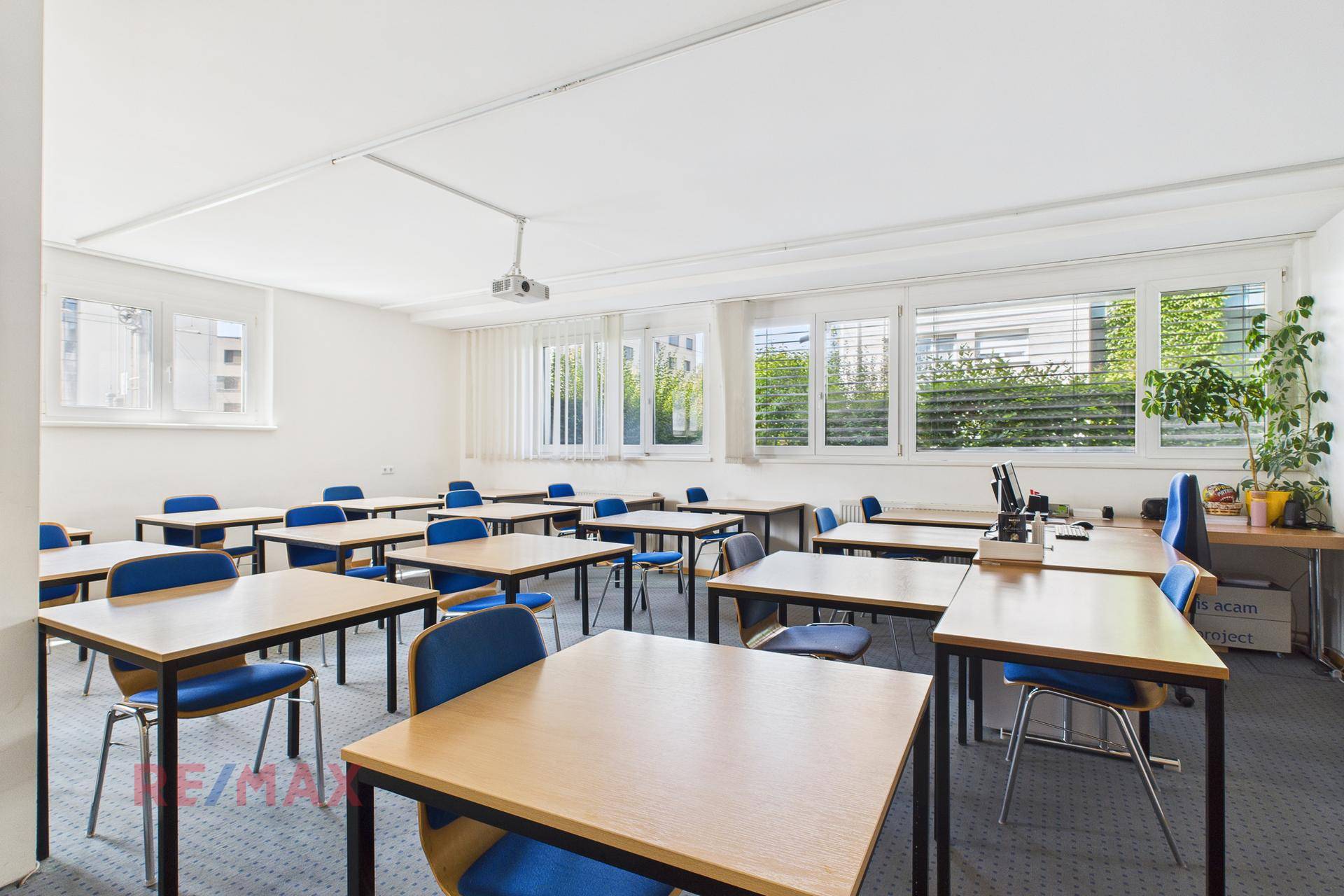 Klassenzimmer mit mehreren Tischen und Stühlen, großen Fenstern und einem Projektor an der Decke.