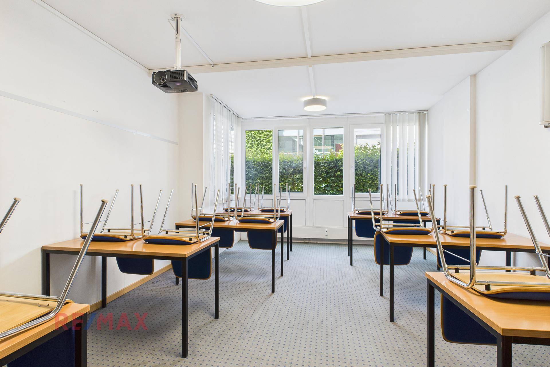 Klassenzimmer mit aufgestellten Tischen und Stühlen, großen Fenstern und hellem Teppichboden.