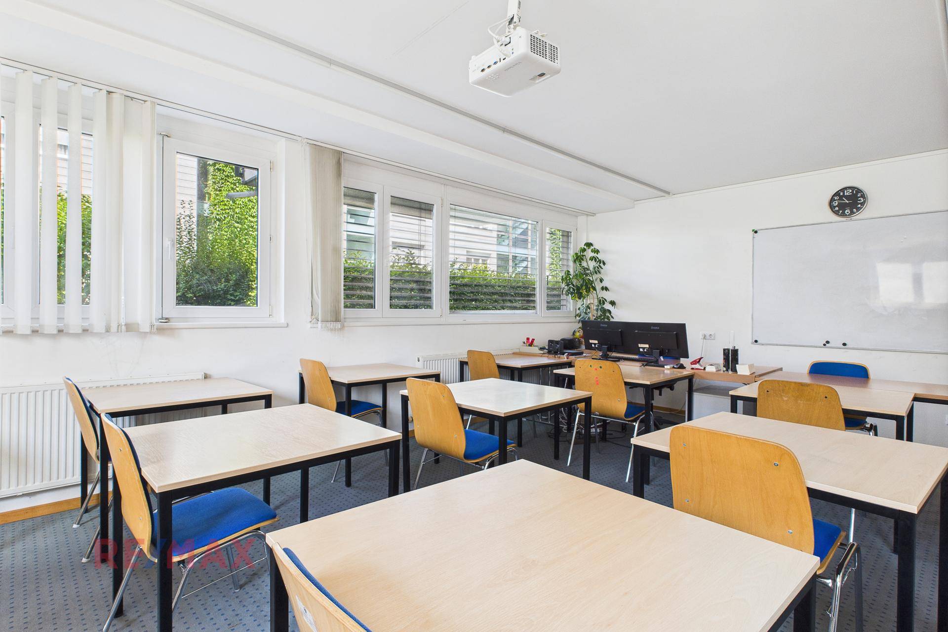 Klassenzimmer mit Tischen und Stühlen, einer weißen Tafel und großen Fenstern mit Jalousien.